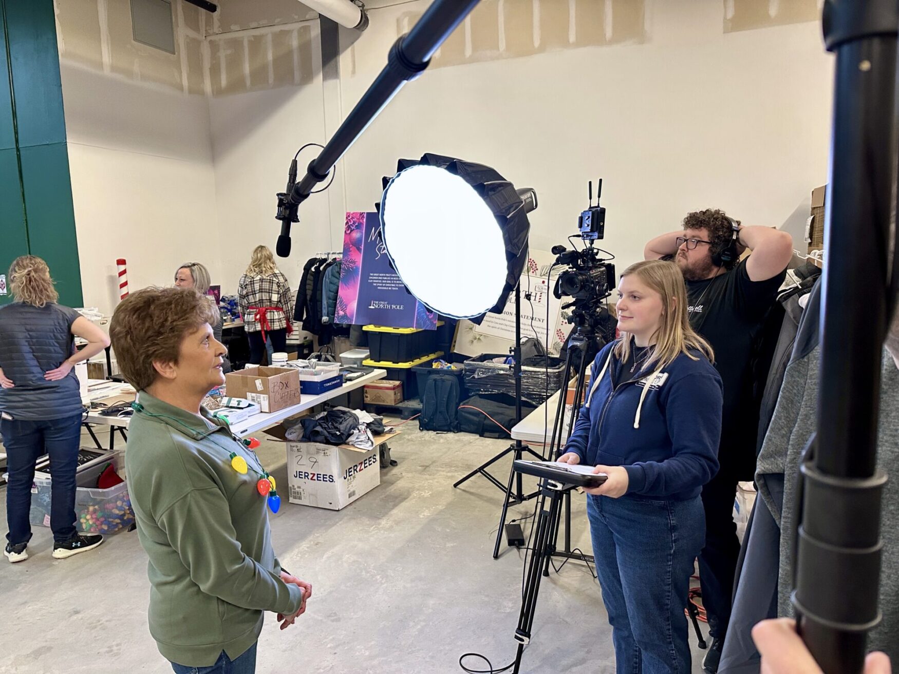 The image shows a behind-the-scenes video shoot. A woman in a green shirt with colorful holiday lights is being interviewed under a bright circular light by someone holding a tablet. A cameraman adjusts his equipment in the background, while others prepare materials in a workshop-like space with holiday decorations and boxes labeled "Jerzees."