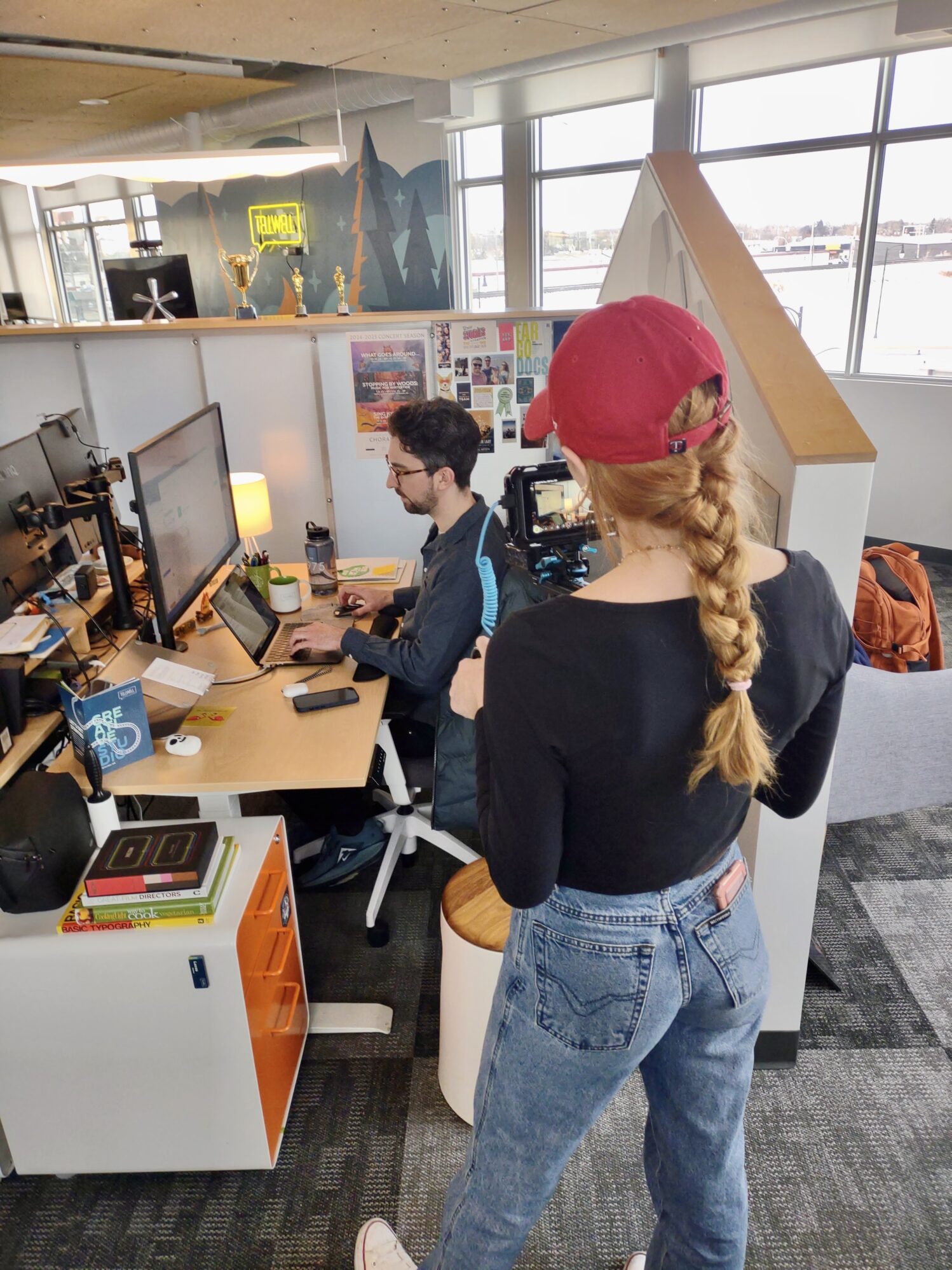 A person with a red hat and braided hair films over the shoulder of another individual working at a desk in a modern office. The desk is decorated with colorful books, posters, and awards, with large windows providing natural light in the background. A "Tellwell" sign is visible on the wall above.