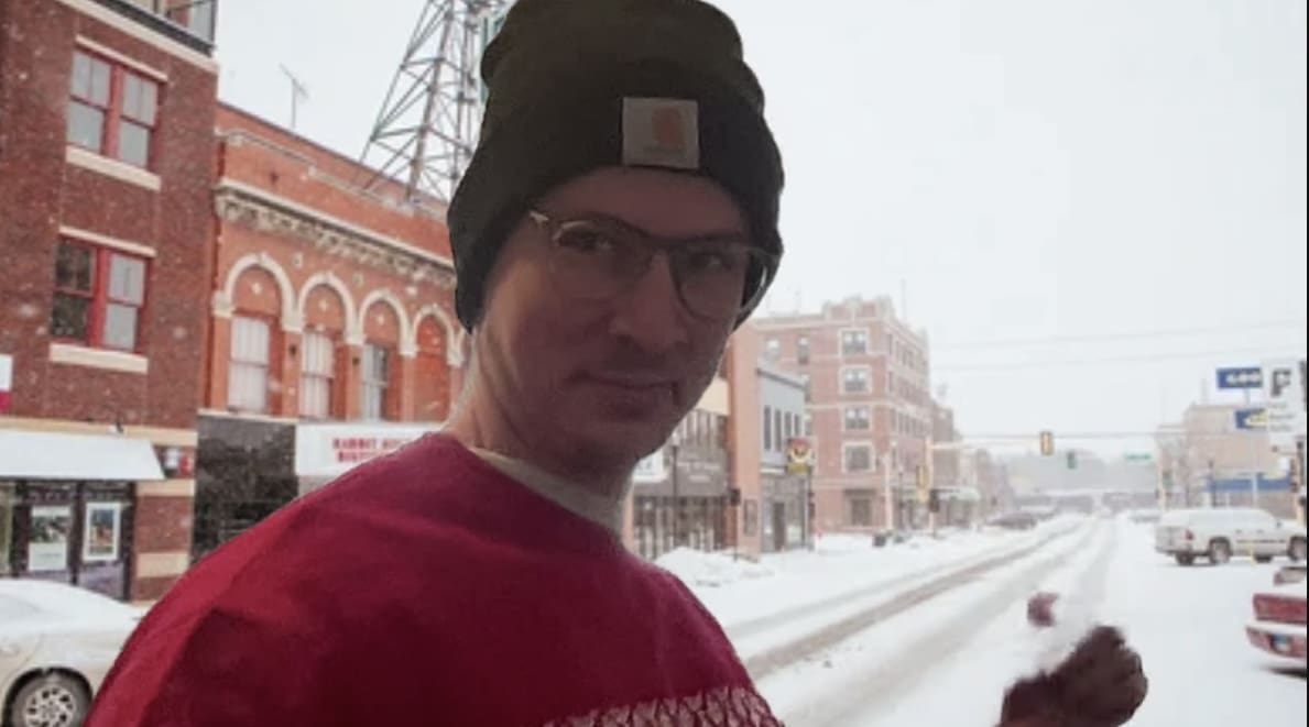 The image shows a man in a red sweater and winter hat in front of a virtual background depicting a snowy downtown street in Fargo.