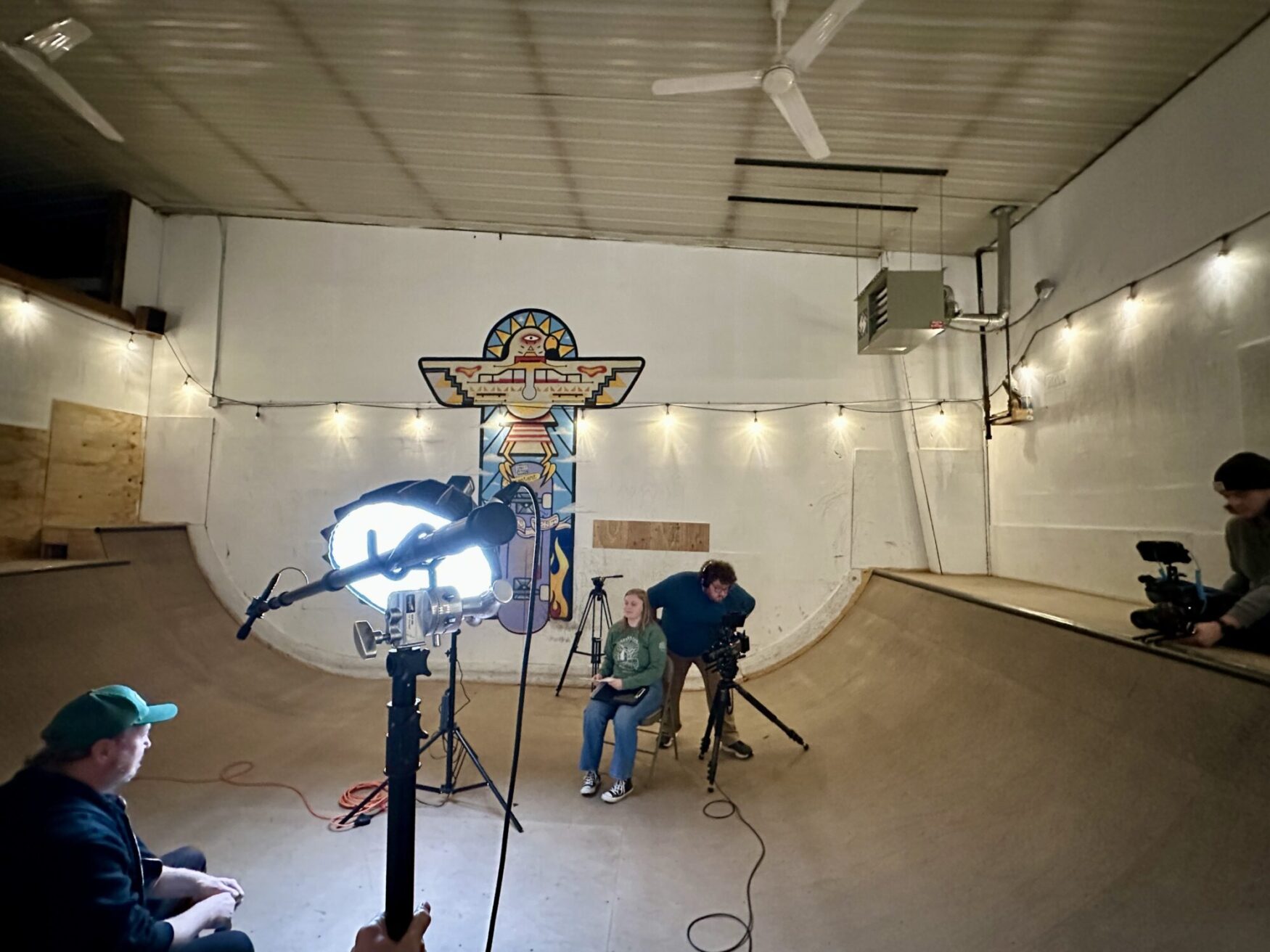 The image shows a filming setup inside a skate park with a ramp on either side. A person sits on a chair in the center, illuminated by a bright key light. Behind them is a colorful totem-inspired mural. Crew members with cameras are positioned around, adjusting equipment, while string lights add a warm, ambient glow to the scene.