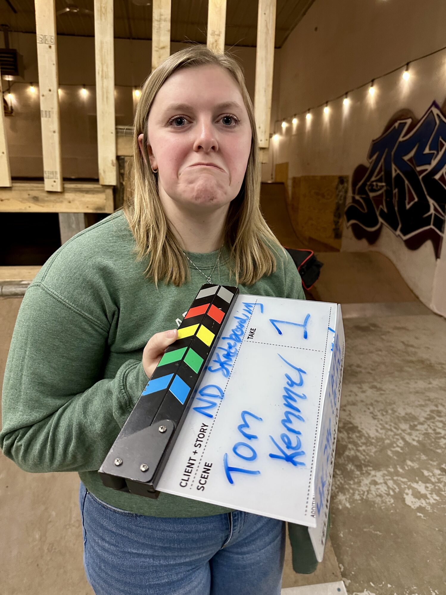 A person in a green sweatshirt holds a film clapperboard labeled "ND Skateboarding" with the name "Tom Kemmer" and "Take 1" written on it. They make a playful, exaggerated frown while standing in a skate park, with string lights and graffiti visible in the background.