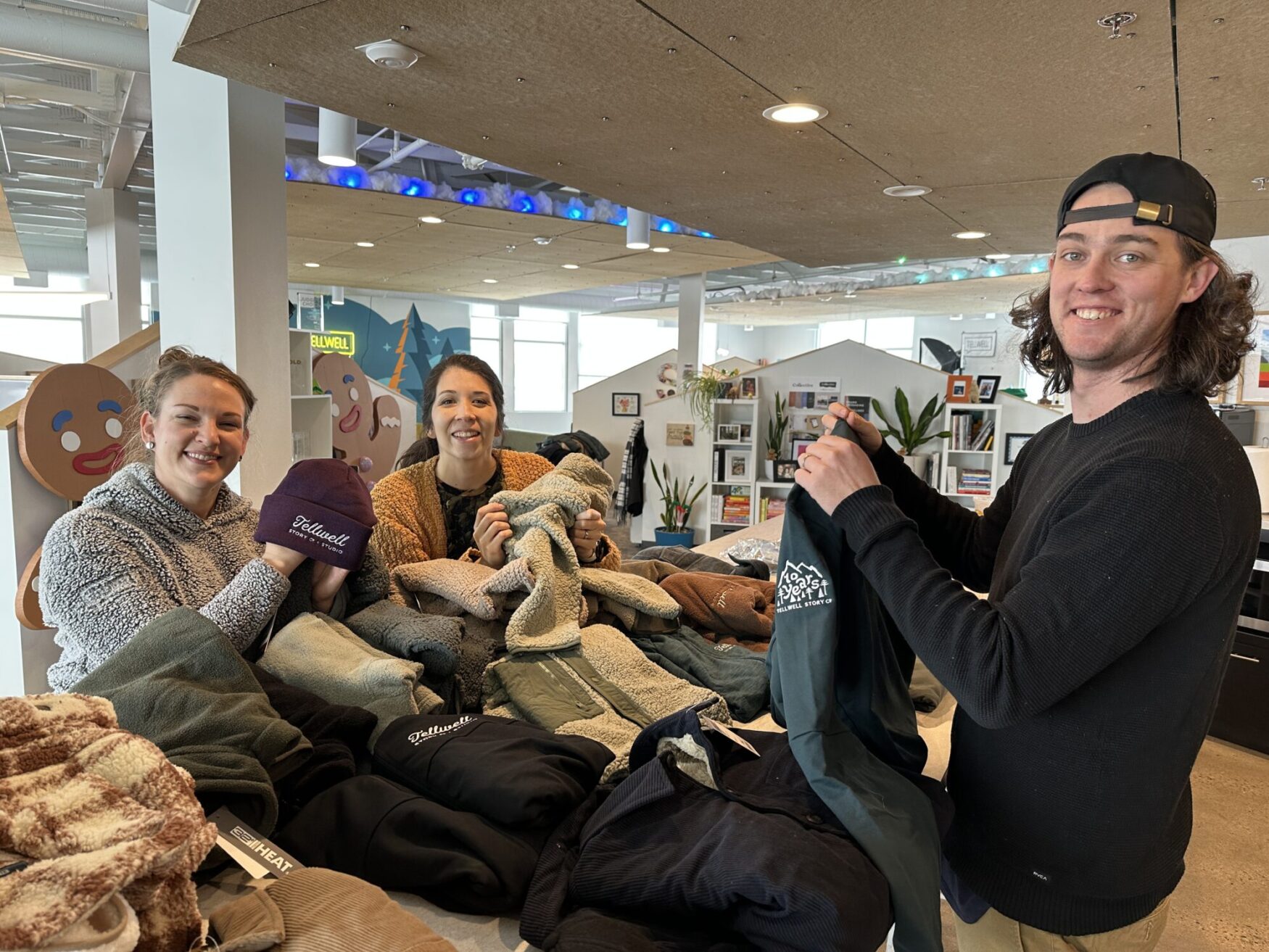 The photo portrays three smiling individuals gathered around a table that is stacked with coats and outerwear.
