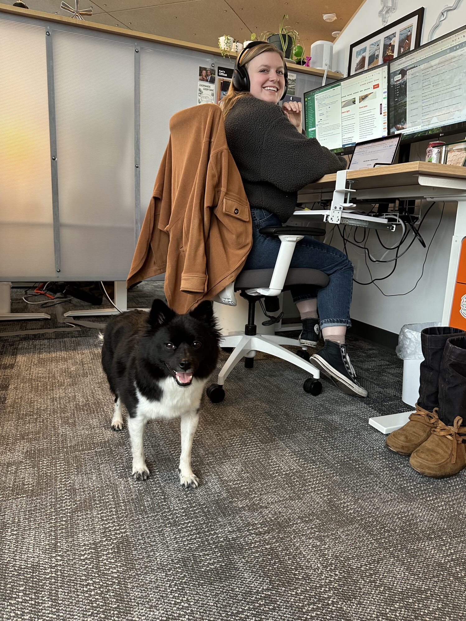 The photo shows a black and white dog looking at the camera in the foreground while a smiling woman looks over her shoulder in the background. The dog is mostly black with white legs and paws.