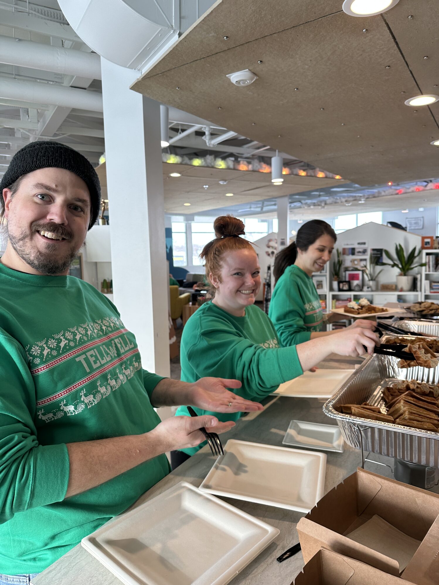 The image shows three people smiling at the camera while they serve breakfast food from a large buffet. They are wearing matching green sweaters that read “Tellwell” across the chest.