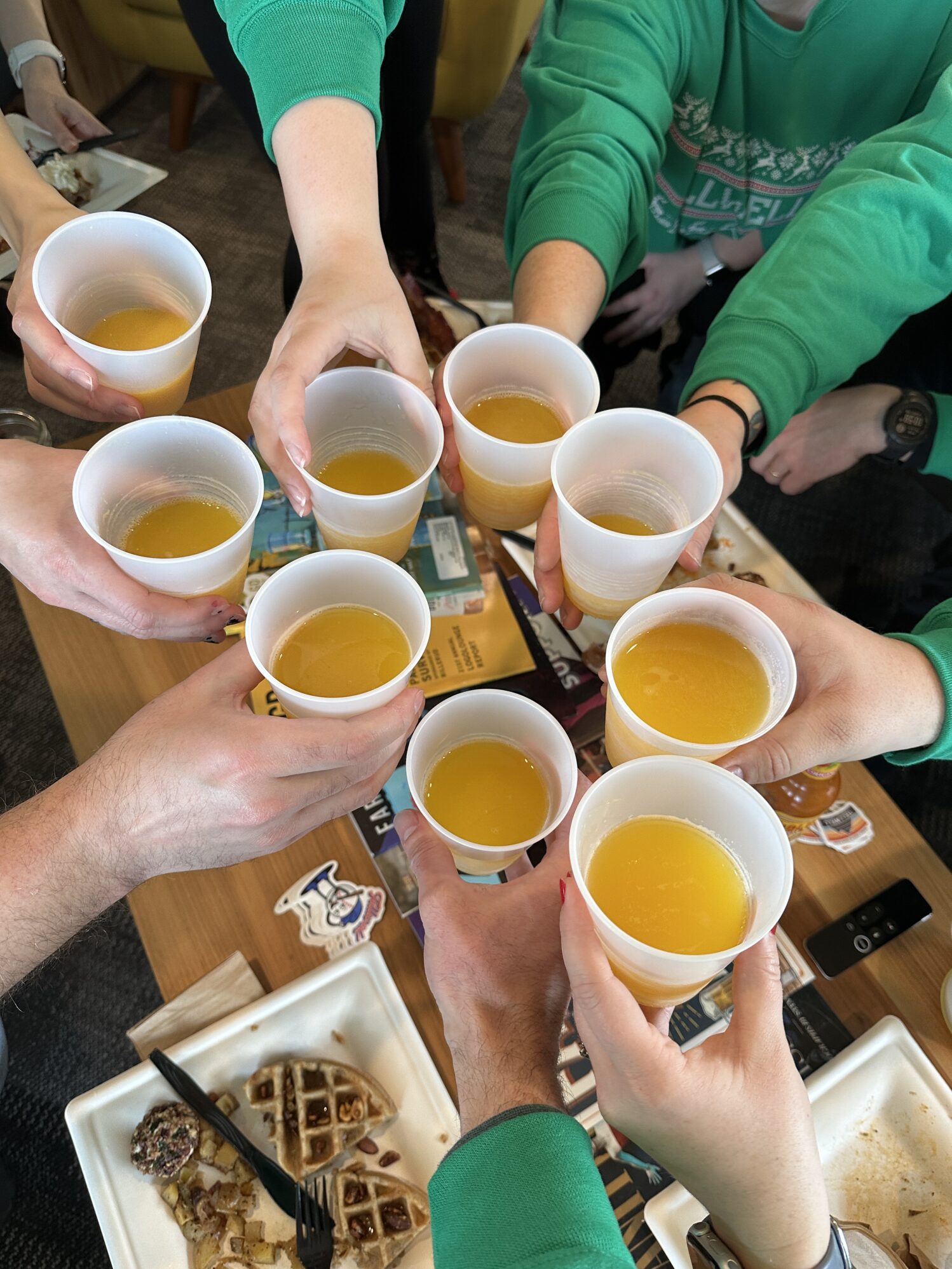 The image shows several plastic cups of orange liquid being brought together by a table full of people.