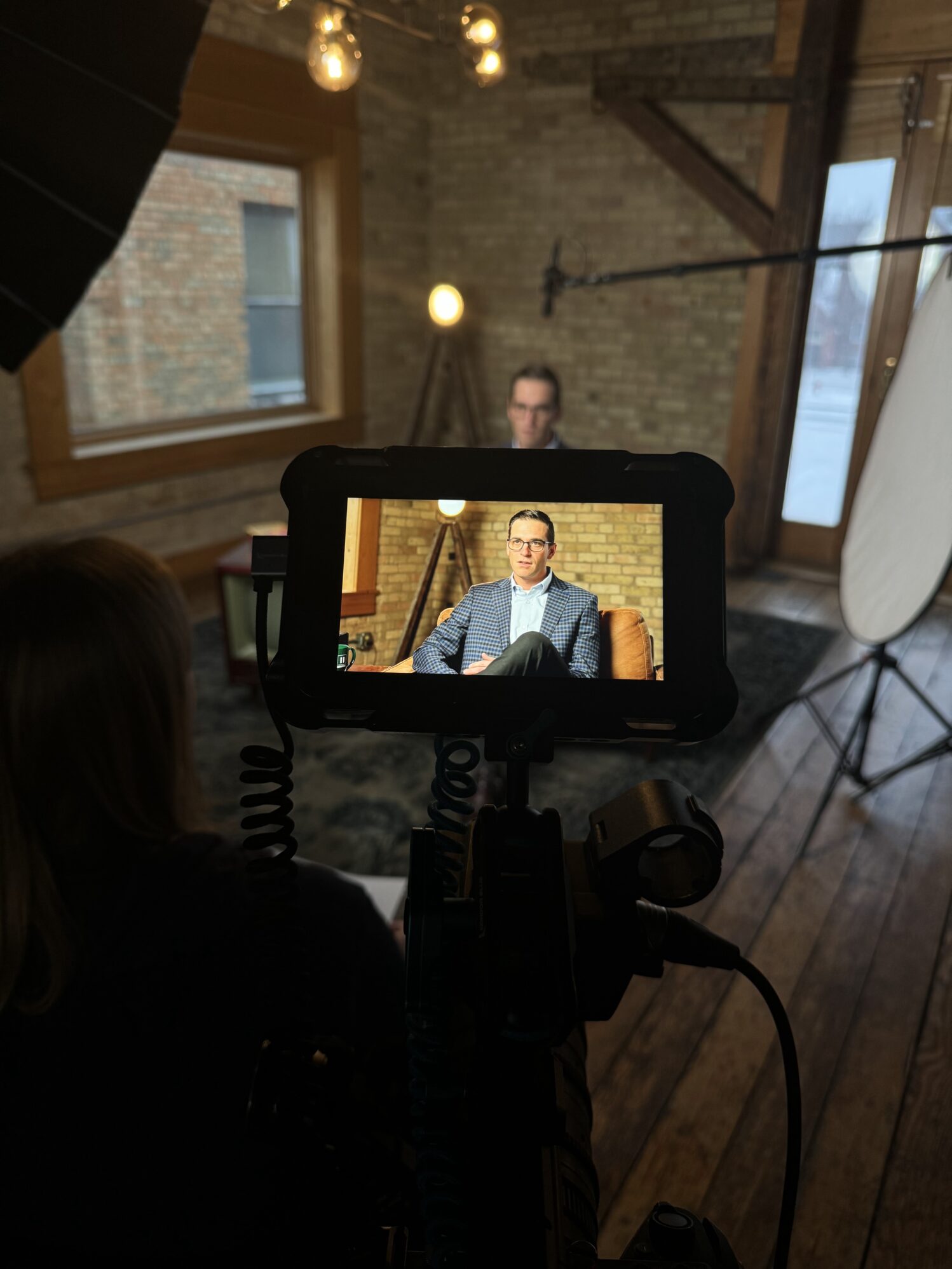 Video interview setup showing a seated man in a warm home with brick walls. Professional filming equipment, including cameras, monitors, a boom mic, and softbox lighting, is prominently displayed in the foreground.