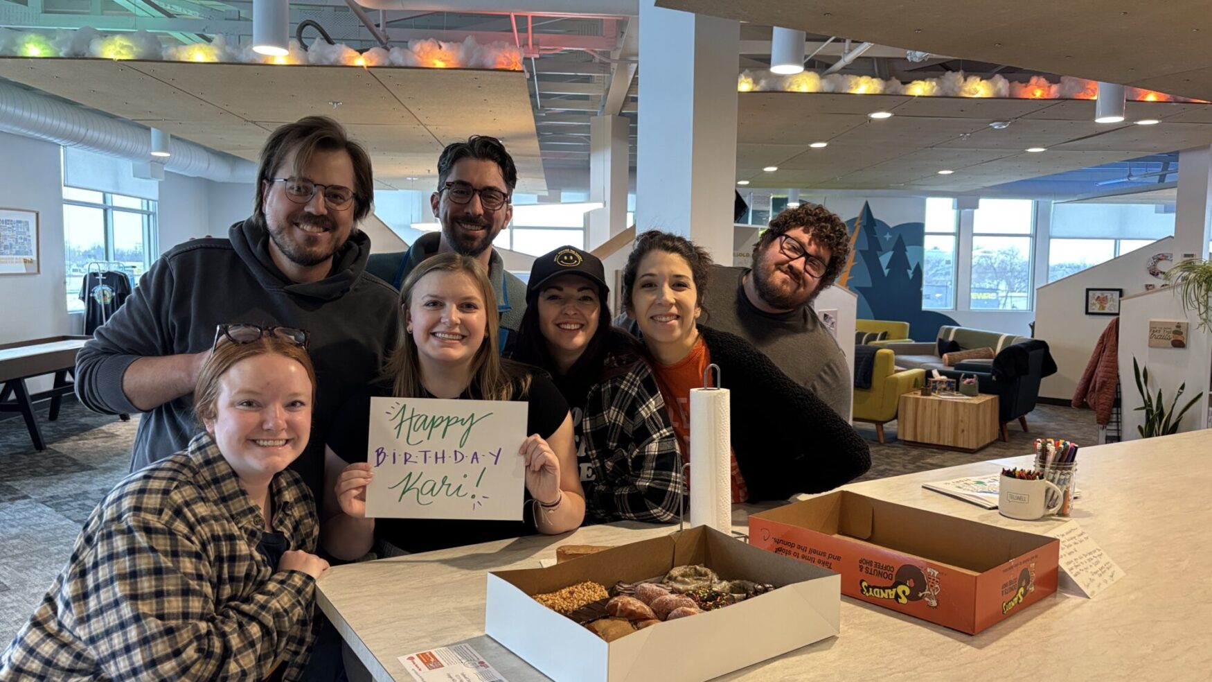Seven people are huddled around a kitchen table, smiling for a photo. One woman is holding a sign that says “Happy birthday Kari!.” There is a box of donuts on the table. They are in a friendly office setting.
