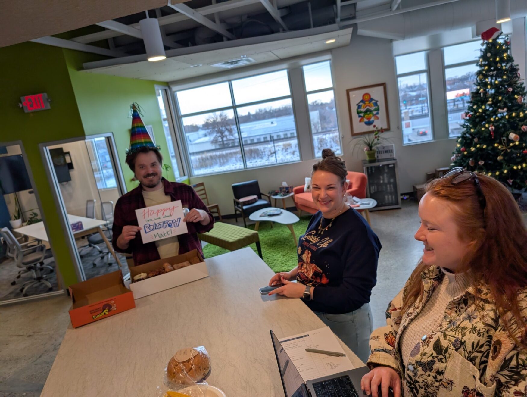 The photo shows three smiling people gathered around a kitchen counter. One of the people is holding a sign that reads “Happy Birthday, Matt”. He is also wearing a colorful cone-shaped birthday hat.