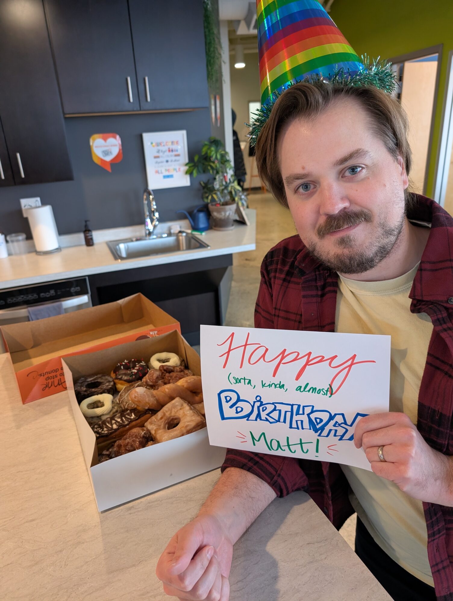 The photo portrays a man leaning on a counter holding a sign that reads “Happy Birthday, Matt”. An open box full of various donuts is resting on the counter.