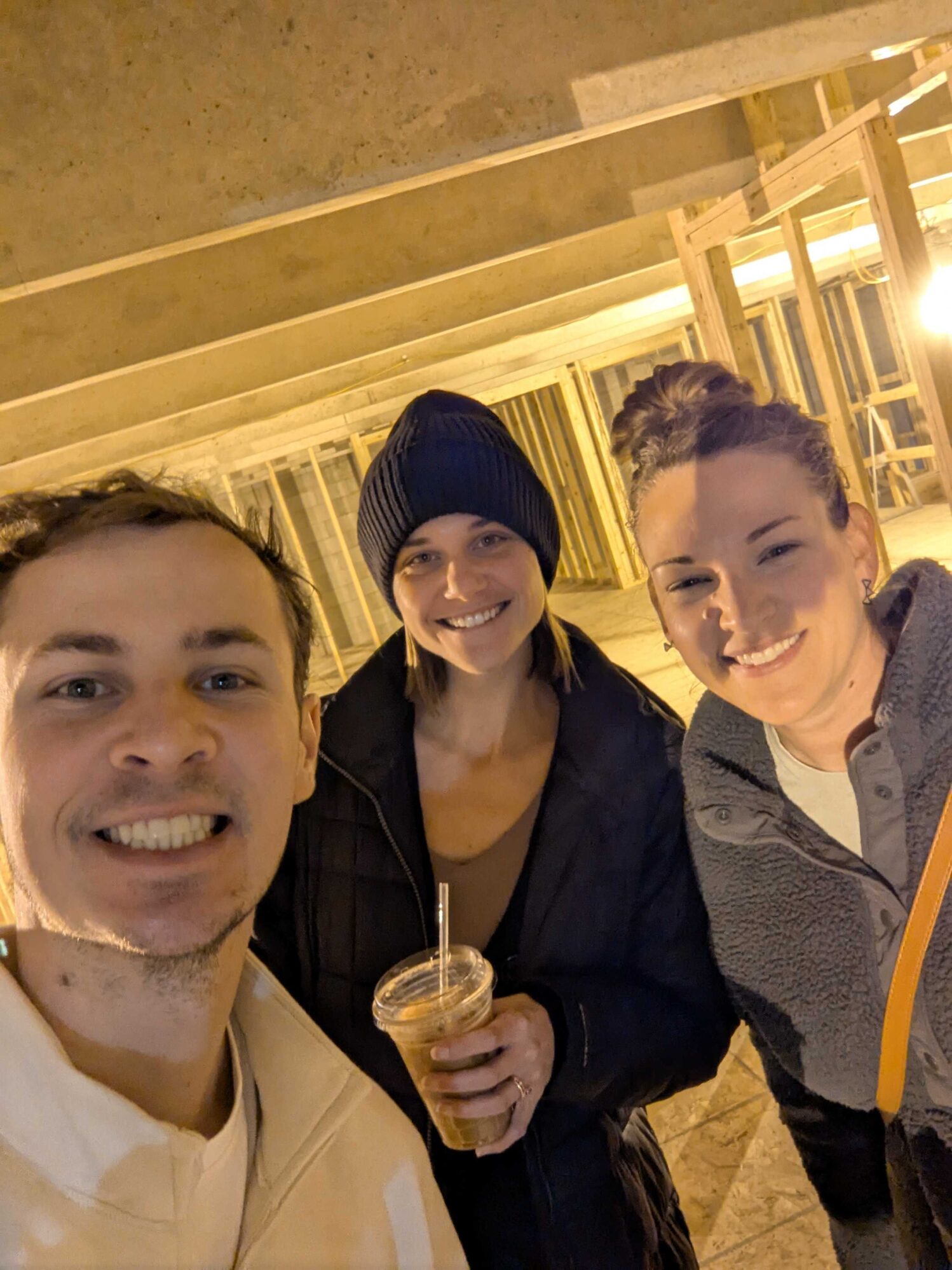 Three people - one man and two women - pose for a selfie in a construction zone. There are beams of wood in the background. One woman is holding an iced coffee.