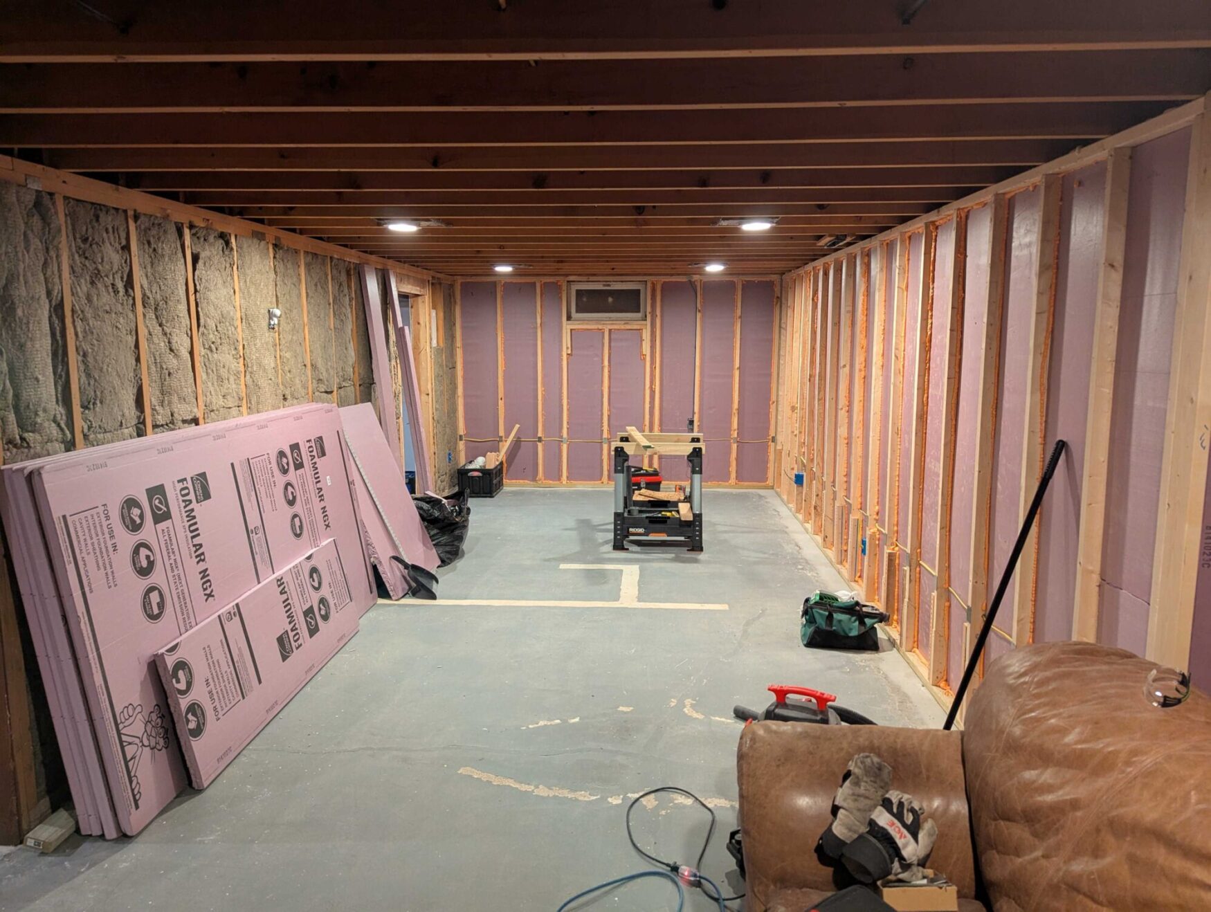 A room under construction. There are structural wooden beams throughout the room, with pink insulation sheets leaning against the wall. There are power tools throughout the room.