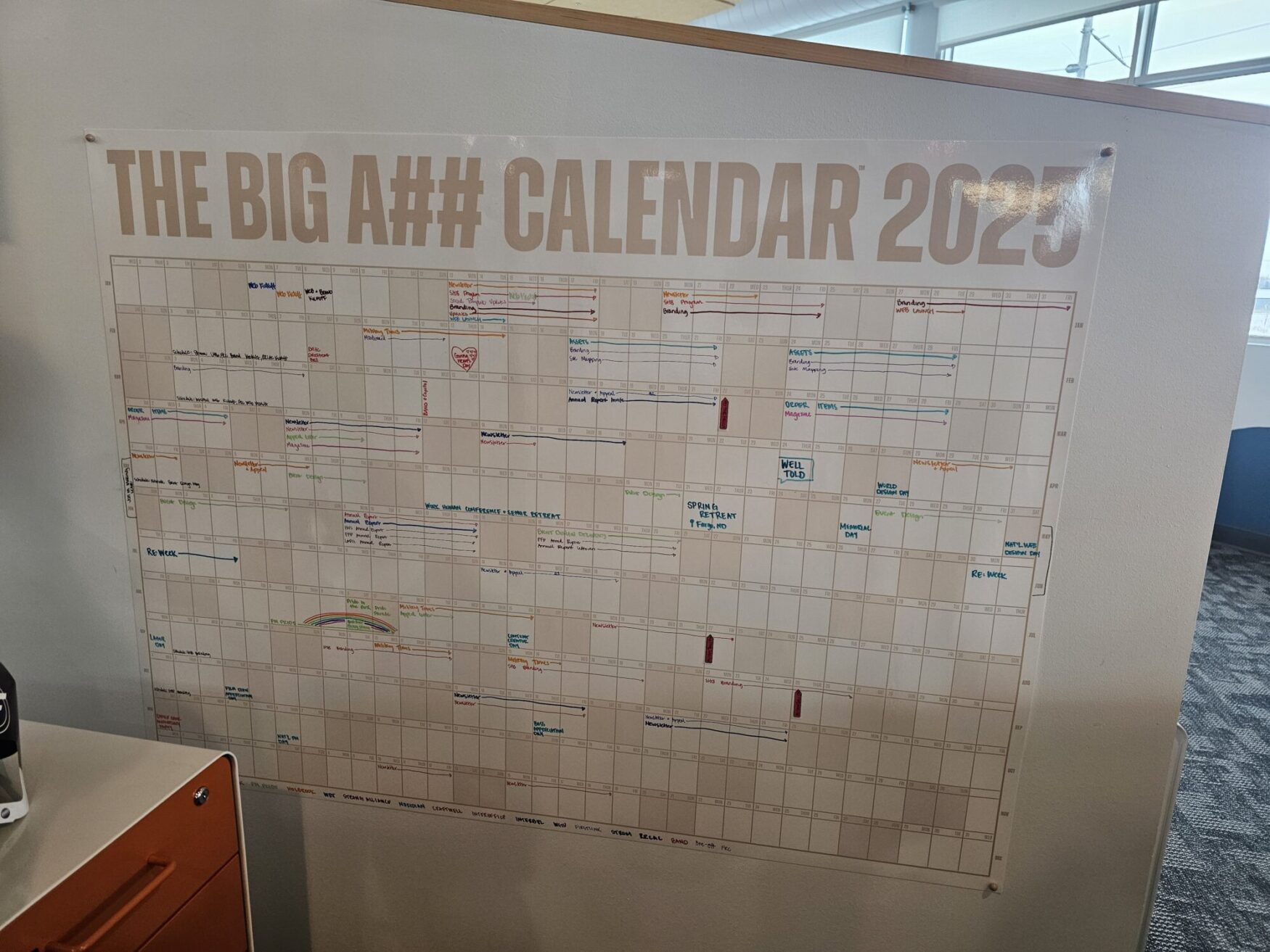 A large wall calendar titled "The Big A## Calendar 2025" displayed in an office setting. The calendar is filled with handwritten events, appointments, and colorful notes spanning all months of the year. The surrounding environment includes a partial view of an orange filing cabinet and a modern office interior with natural light streaming in from the windows.