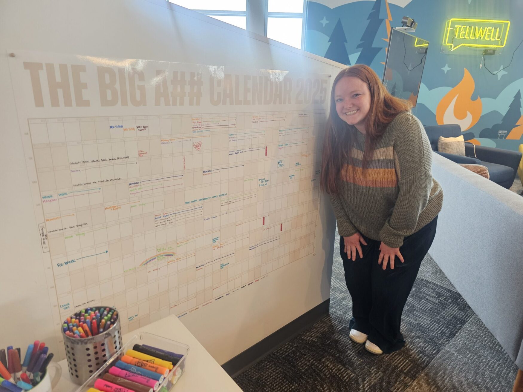 A smiling woman with long red hair, wearing a green sweater with horizontal stripes and black pants, is crouched slightly beside a large wall calendar titled "THE BIG A## CALENDAR 2025." The calendar is filled with colorful notes and markings. In front of the calendar is a table holding containers of colorful markers and pens. The background features a bright, cheerful office space with a neon "TELLWELL" sign, a blue wall with stylized trees and a campfire illustration, and comfortable seating.