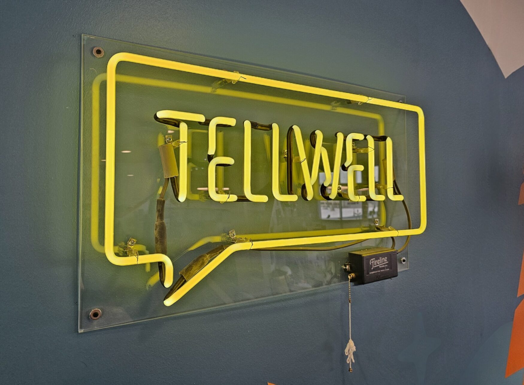 The image shows a bright yellow neon sign reading 'TELLWELL' in a speech bubble shape, mounted on a teal wall with visible wiring and a Fireline switch box attached.