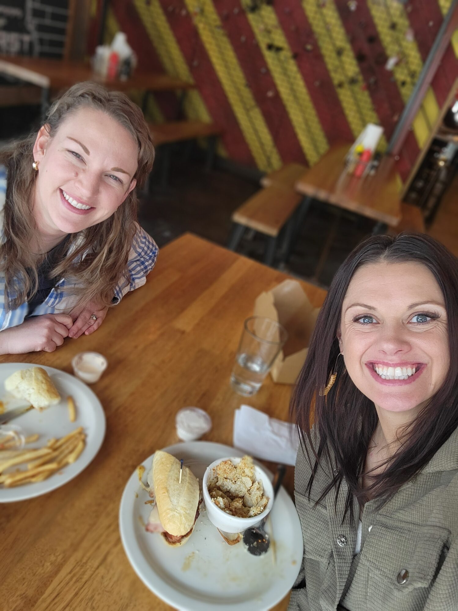 Two women smiling at the camera while seated at a wooden table in a restaurant. Plates with sandwiches, fries, and a bowl of food are visible in front of them. The background features colorful red and yellow diagonal stripes and bench seating.