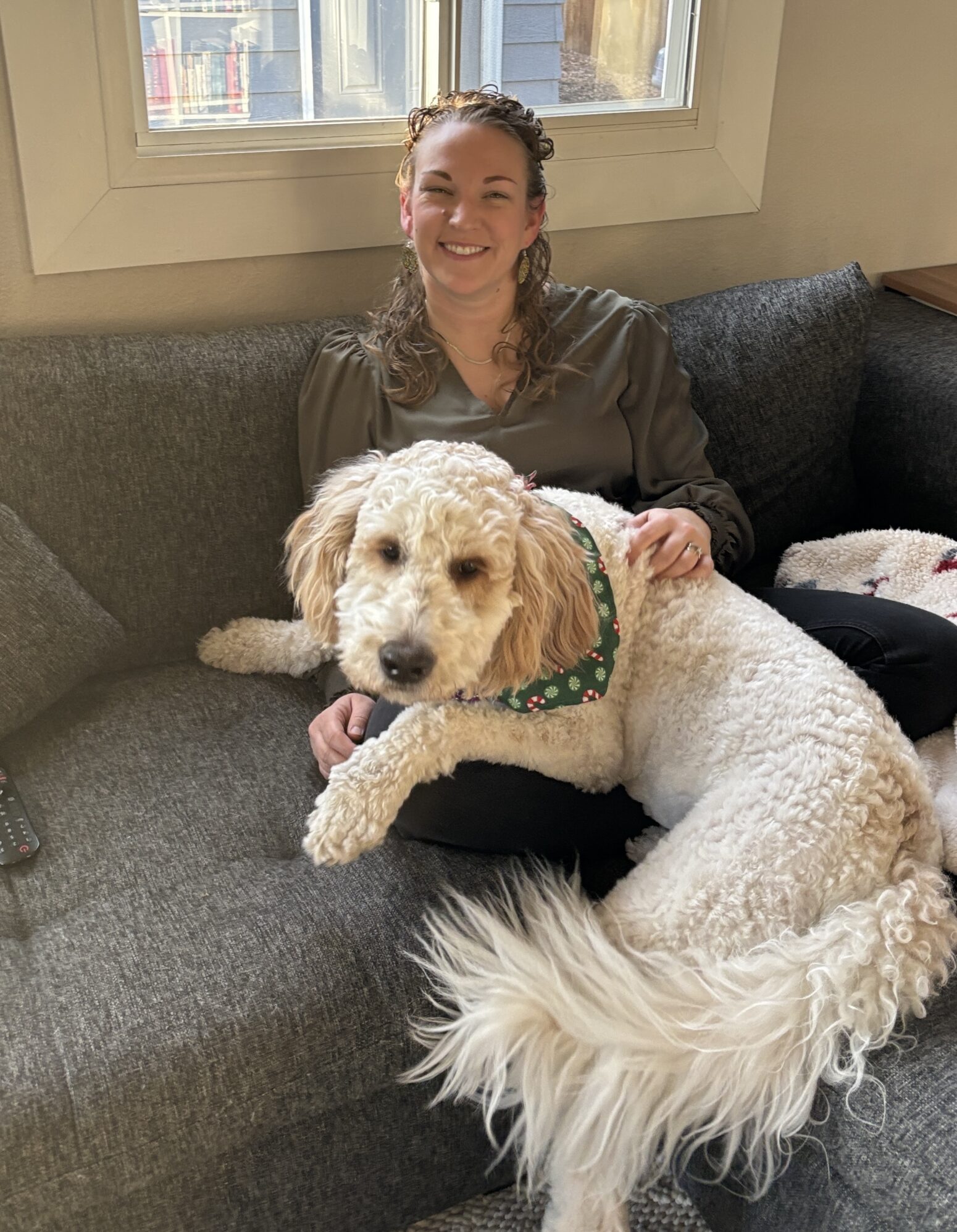 A smiling person sits on a gray couch in a cozy room with a large window behind them. A fluffy white dog with a festive green bandana lies comfortably across their lap. The window shows a backyard with trees and a shed.