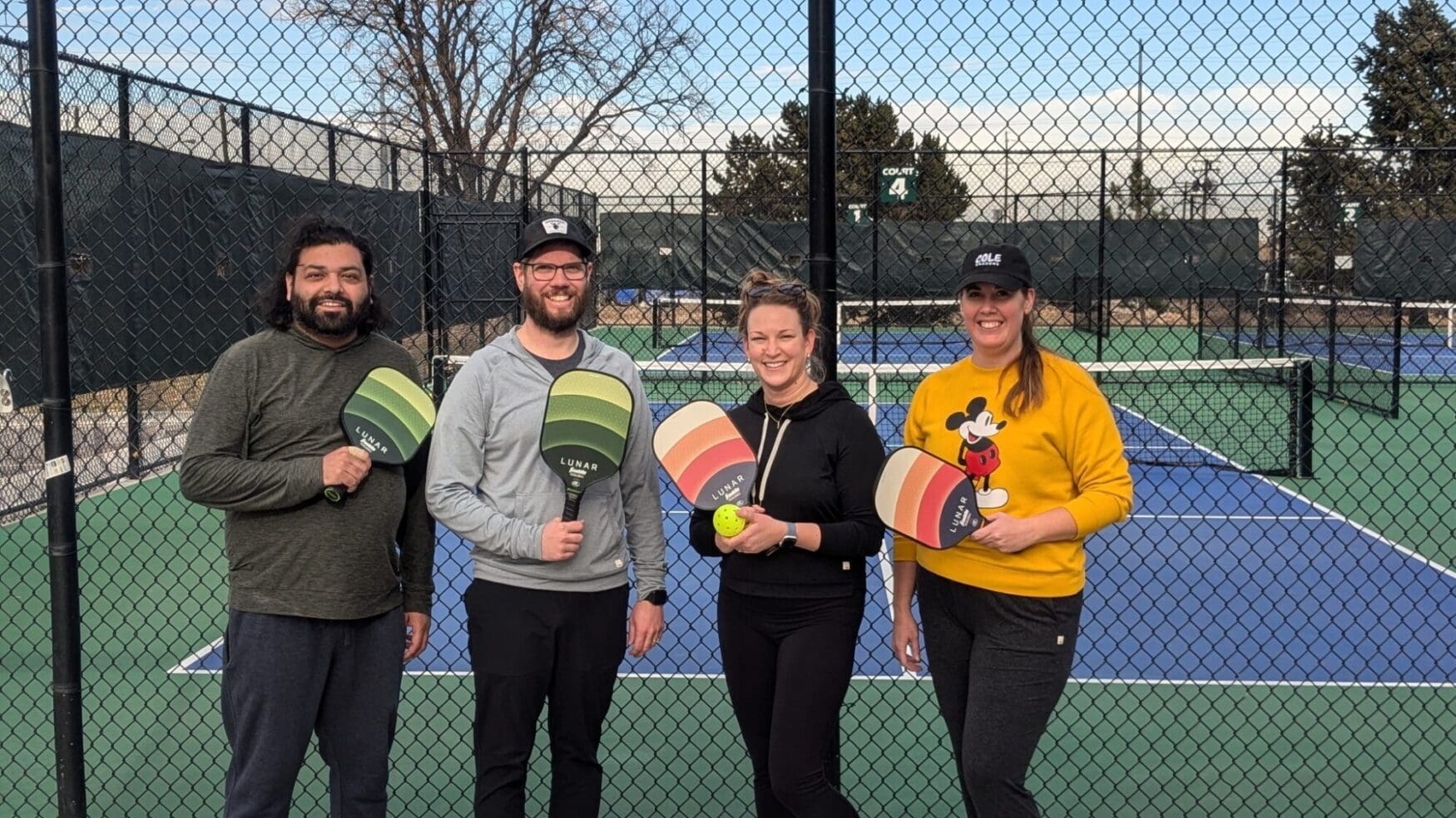A group of four people standing on an outdoor pickleball court, each holding a paddle and smiling. Two men and two women pose in front of the court's chain-link fence, with blue and green courts visible in the background. One person wears a Mickey Mouse sweater, and another has a pickleball in hand.