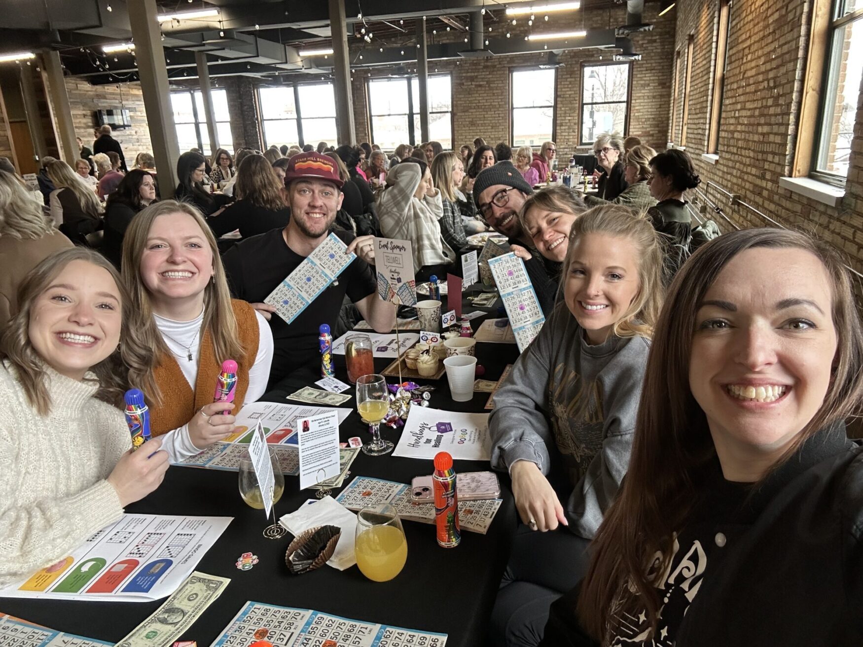 A group of seven smiling people sits at a long table covered with bingo cards, colorful daubers, drinks, and small decorations. They are in a lively venue with exposed brick walls, large windows, and a crowd of other bingo players in the background. One person holds up a bingo card, while another displays a sign that reads "Event Sponsors: TELLWELL." The atmosphere is fun and social, with everyone appearing excited and engaged in the game.