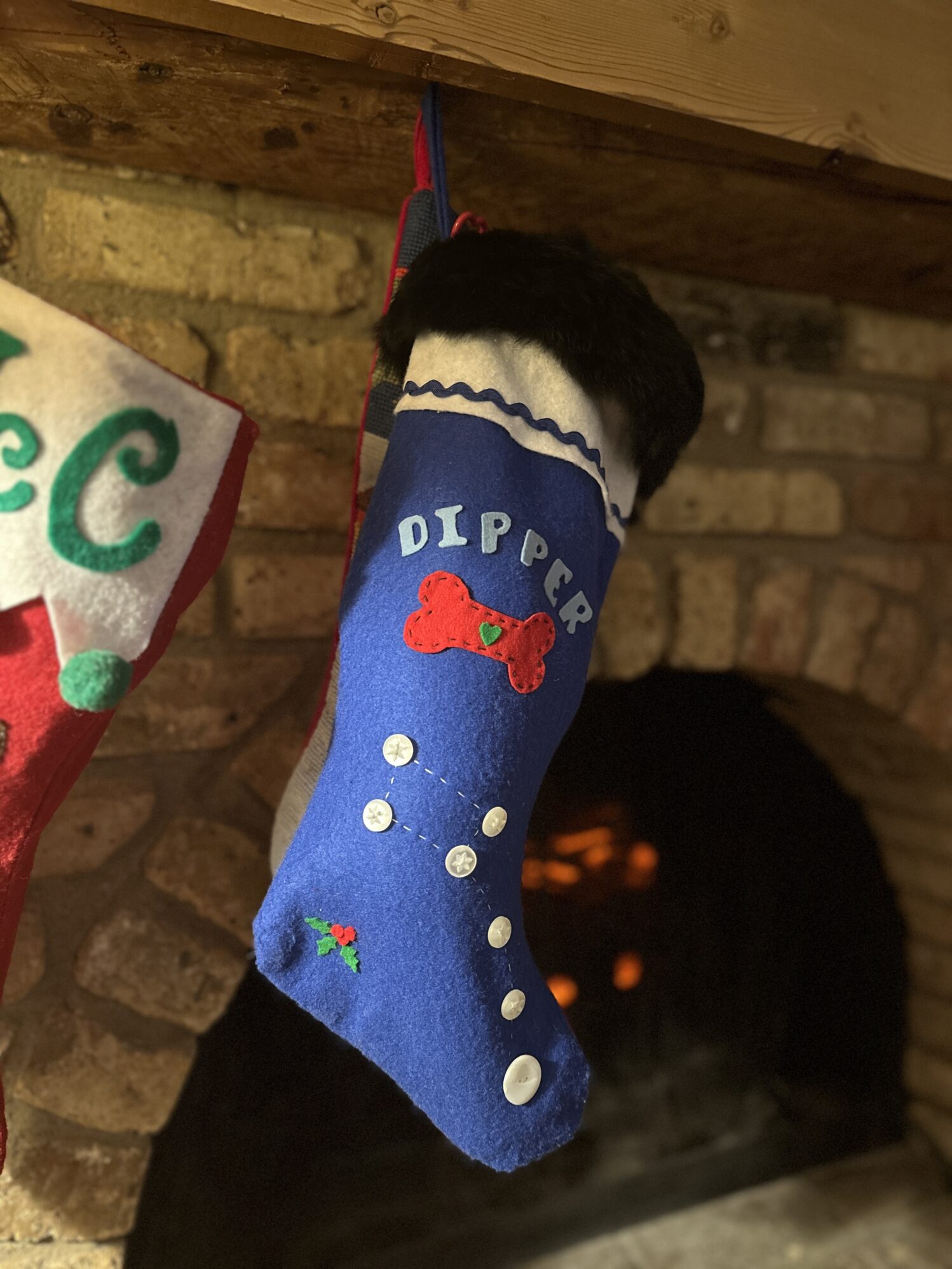 A Christmas stocking hanging on a brick fireplace mantel. The stocking is blue and white with black fur trim. The stocking says “Dipper” and has a red dog bone and the Little Dipper constellation on it, making it appear to be a stocking for a dog.