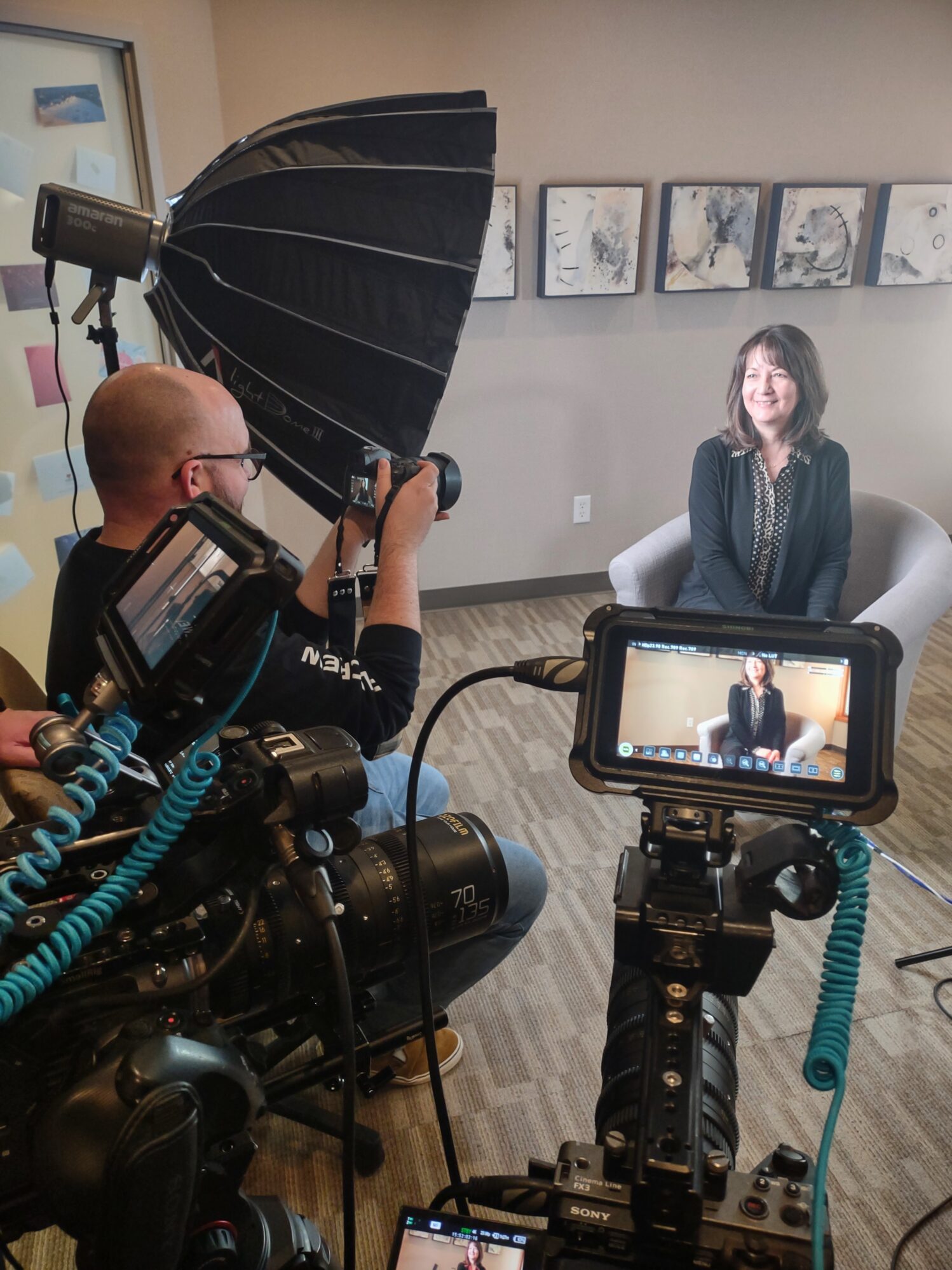 A woman is seated in a modern office setting, smiling as she participates in an on-camera interview. Multiple professional cameras are set up to capture the scene, including a close-up view displayed on a monitor. A photographer operates a camera with a large lens while a softbox light provides even illumination. Abstract artwork hangs on the wall in the background, and the room's neutral tones contribute to a polished and professional atmosphere.