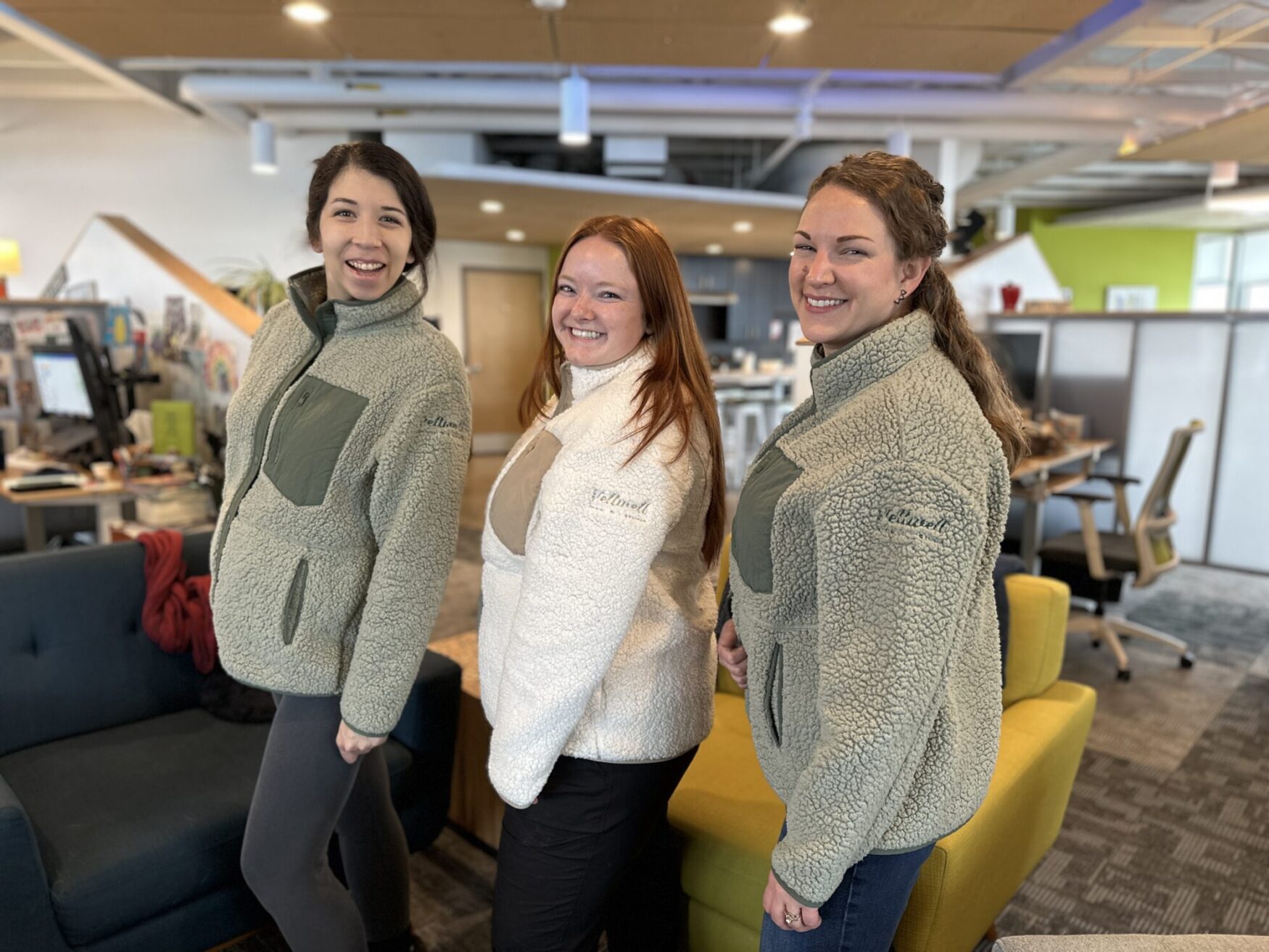 Three smiling people pose in a brightly lit office space, wearing matching sherpa jackets with the "Tellwell" logo embroidered on the sleeves. The office features modern furniture, including a teal couch and yellow chairs, with a background of desks and vibrant decor.