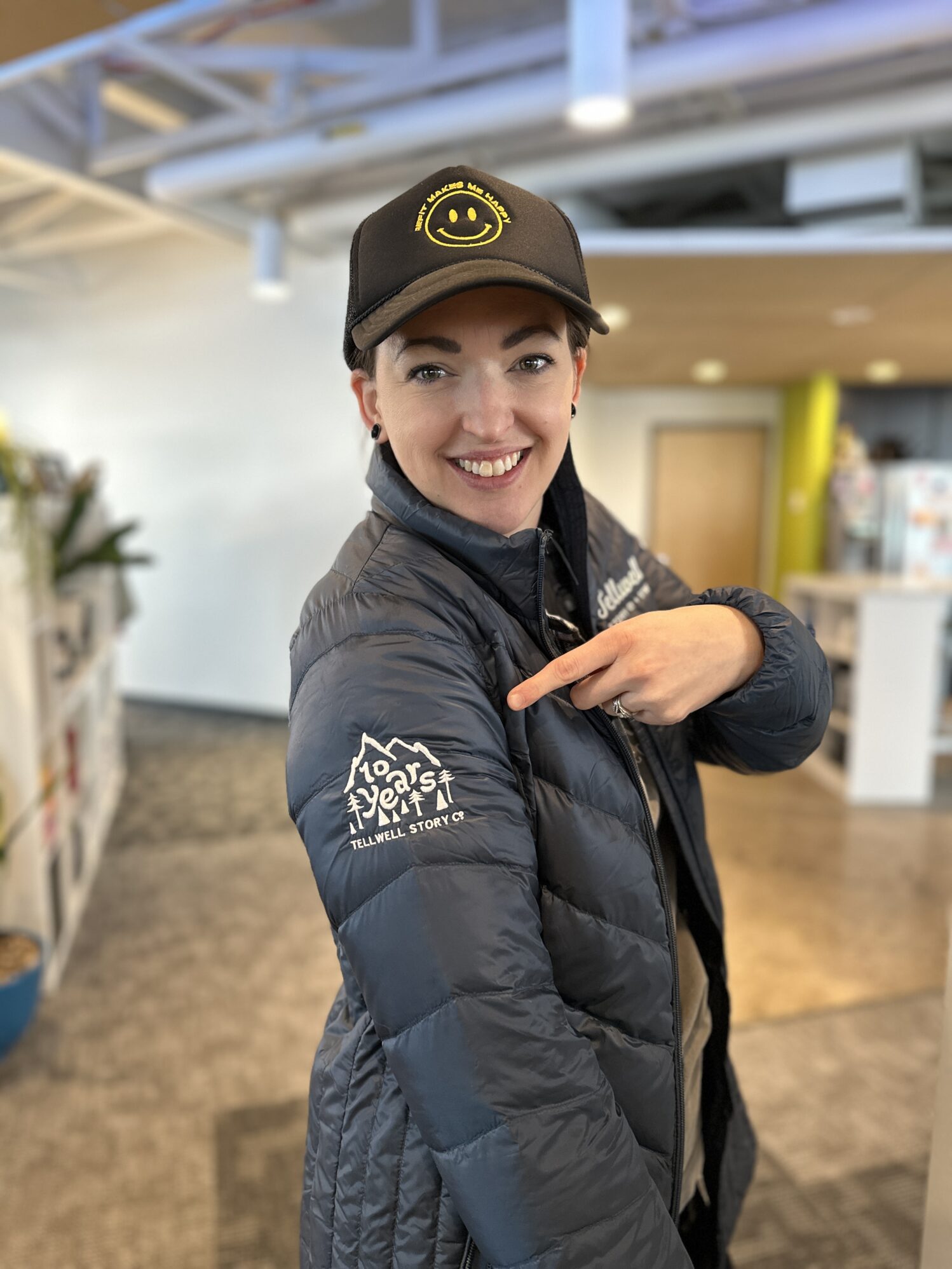 A smiling person wearing a black baseball cap points to their navy blue puffer jacket embroidered with the text "10 Years Tellwell Story Co." They are standing in an office with a bookshelf in the background.