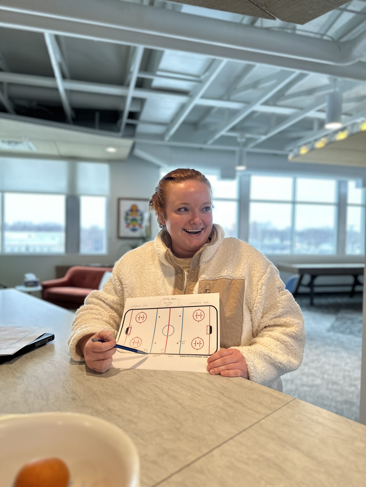 A woman holding a piece of paper showing the layout of a hockey rink. She is pointing at it with a pencil, explaining something on the paper.