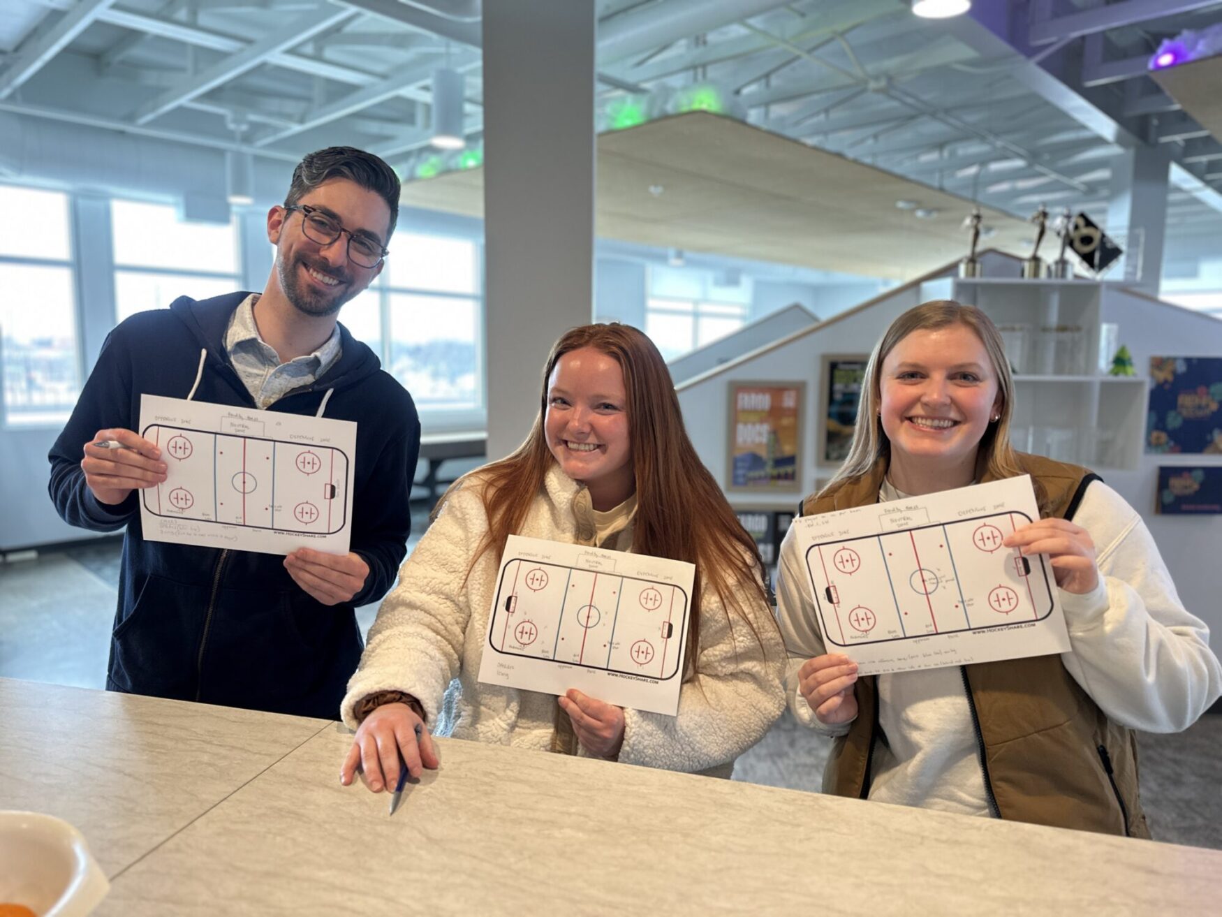 One man and two women pose for a photo, smiling and holding pieces of paper showing the layout of a hockey rink. They are in an office setting.