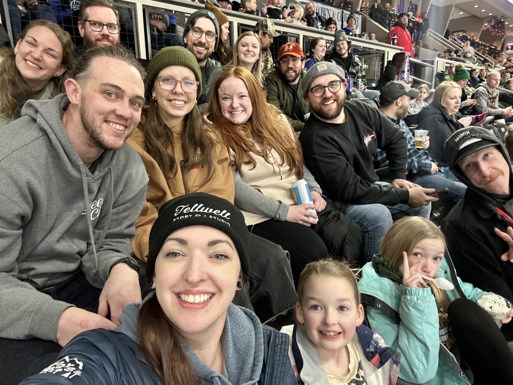 The image shows a lively group photo at a sports event, featuring adults and children sitting together in the stands, smiling and enjoying the atmosphere. The crowd is visible in the background, with some holding drinks and snacks. One person in the front is wearing a black beanie with the 'Tellwell Story Co. & Studio' logo.