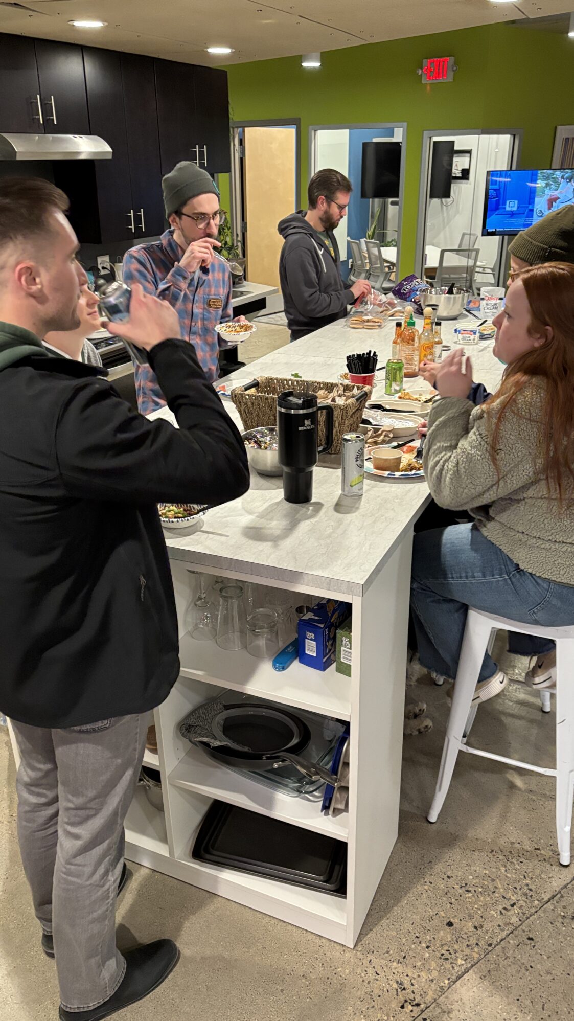 A group of people gathers around a kitchen island, enjoying food and drinks in a casual setting. The countertop is filled with snacks, hot sauce bottles, and drinks, creating a lively and informal atmosphere. In the background, a lime-green wall and an open doorway add a vibrant touch to the space, while a TV is visible in another room.