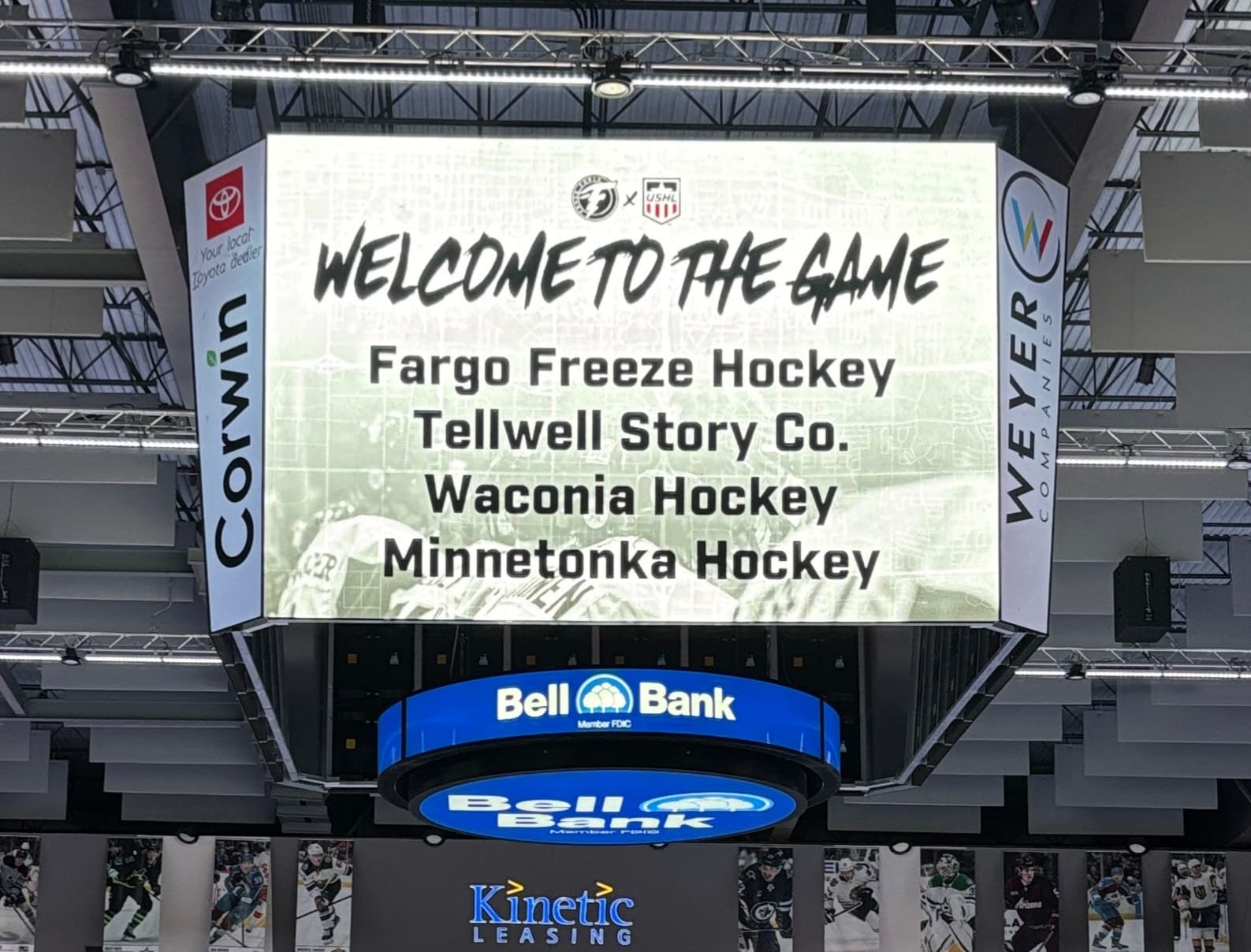 The image shows a digital scoreboard in an arena displaying a message reading 'WELCOME TO THE GAME' with a list of names: Fargo Freeze Hockey, Tellwell Story Co., Waconia Hockey, and Minnetonka Hockey. The scoreboard is surrounded by advertisements, including 'Corwin' and 'Weyer Companies,' and is topped with a Bell Bank logo.