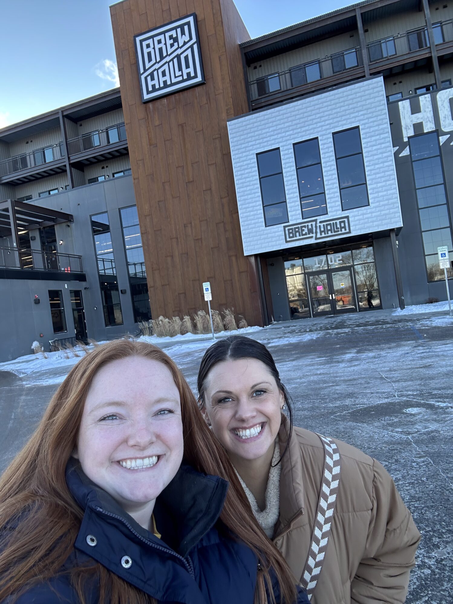 Two women smiling for a selfie outside a building with the sign "Brewhalla" on its façade. The building has a modern design with wooden and metal accents, and the snowy ground indicates a winter setting.