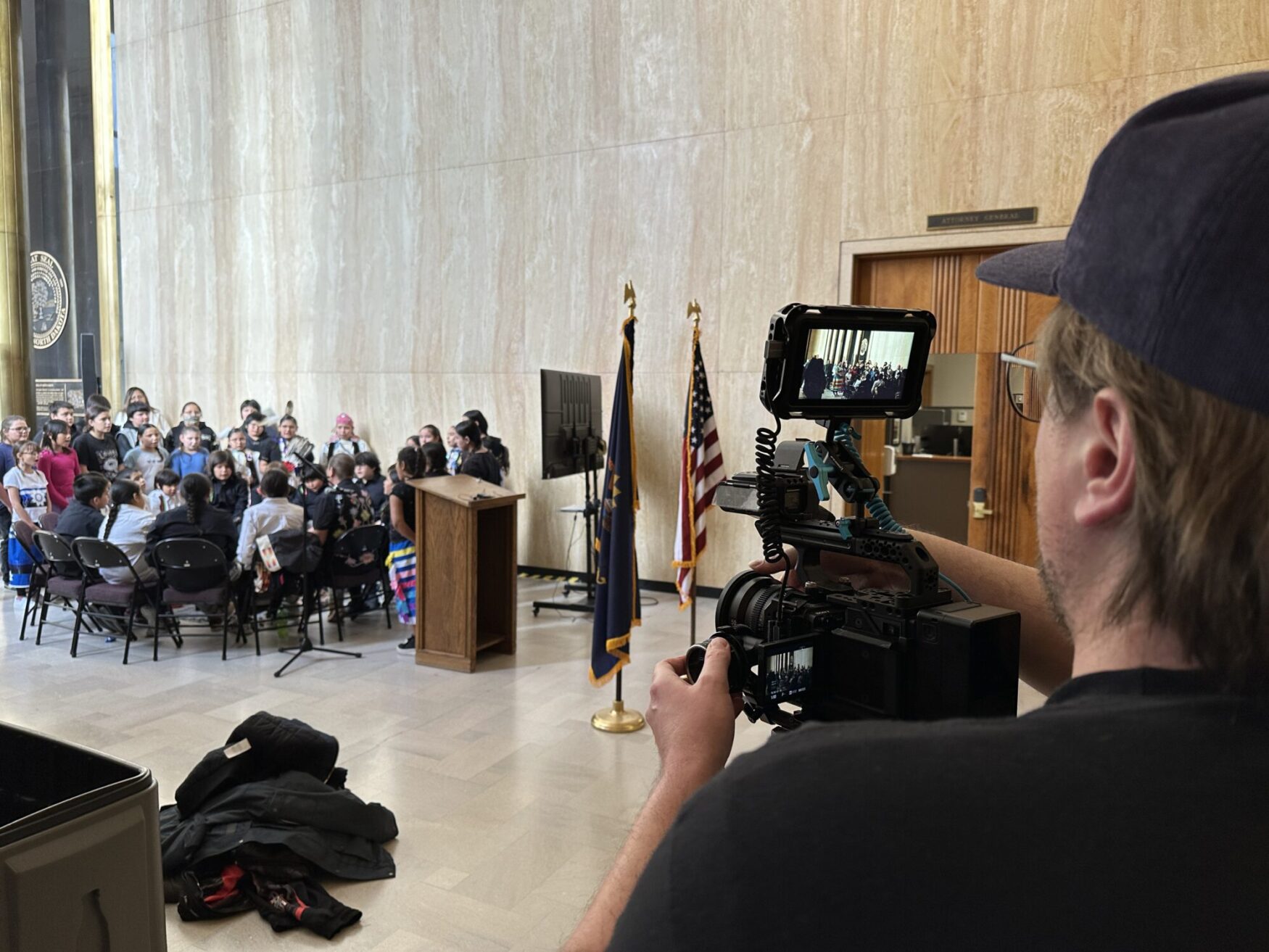 A person wearing a cap operates a professional camera with a monitor, filming a group of children seated and standing in front of a podium in a large hall. The scene includes two flags, one American and one state, as well as a marble wall and wooden doors in the background. The children appear to be part of a formal or celebratory event, with some wearing traditional clothing. The setup suggests an official or cultural gathering, possibly in a government building.