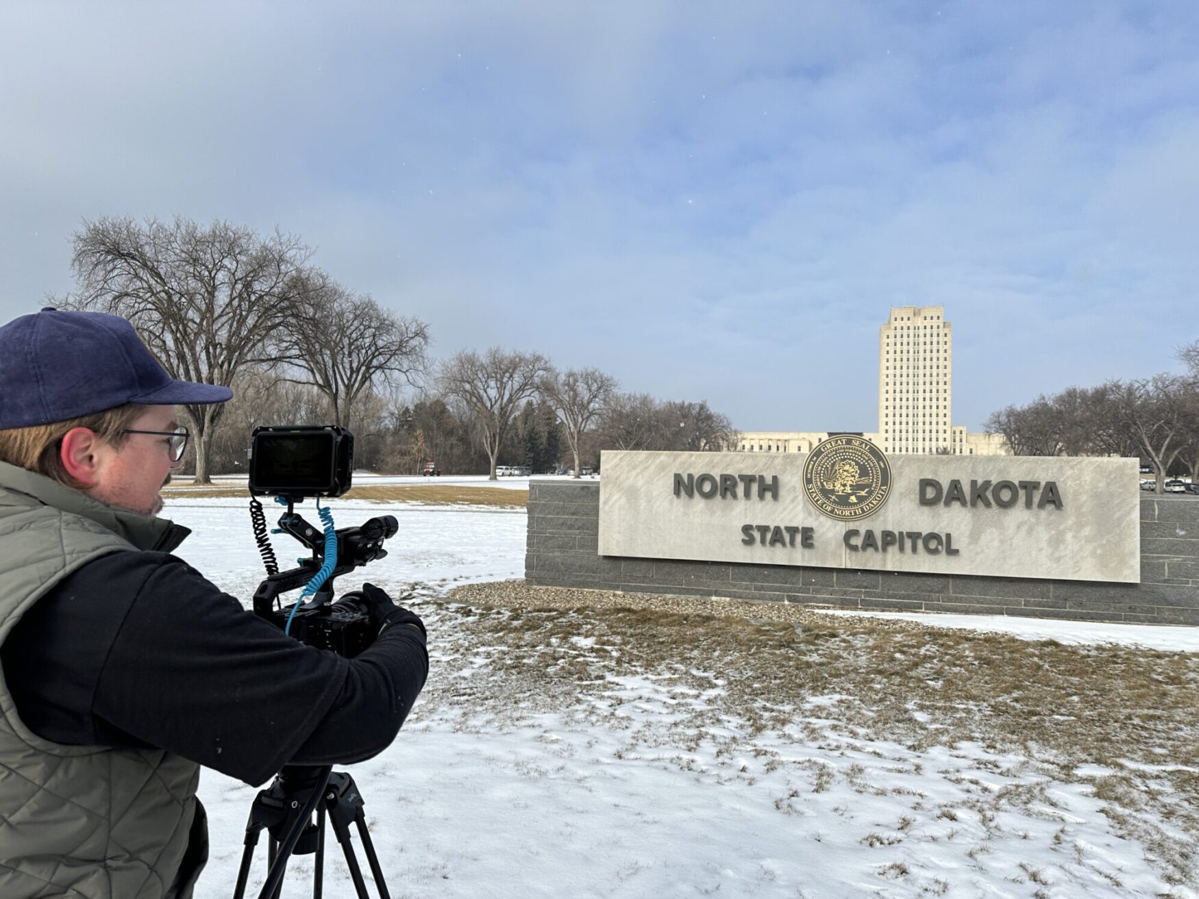 A person wearing a cap operates a professional camera with a monitor, filming outside at a sign that reads “North Dakota State Capitol”. The scene includes snow on the ground, leafless trees, and a capitol building in the background.