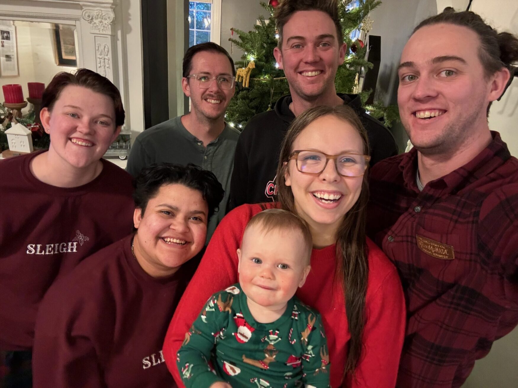 A happy gathering of people smiling for a photo in front of a festive Christmas tree adorned with lights and ornaments.