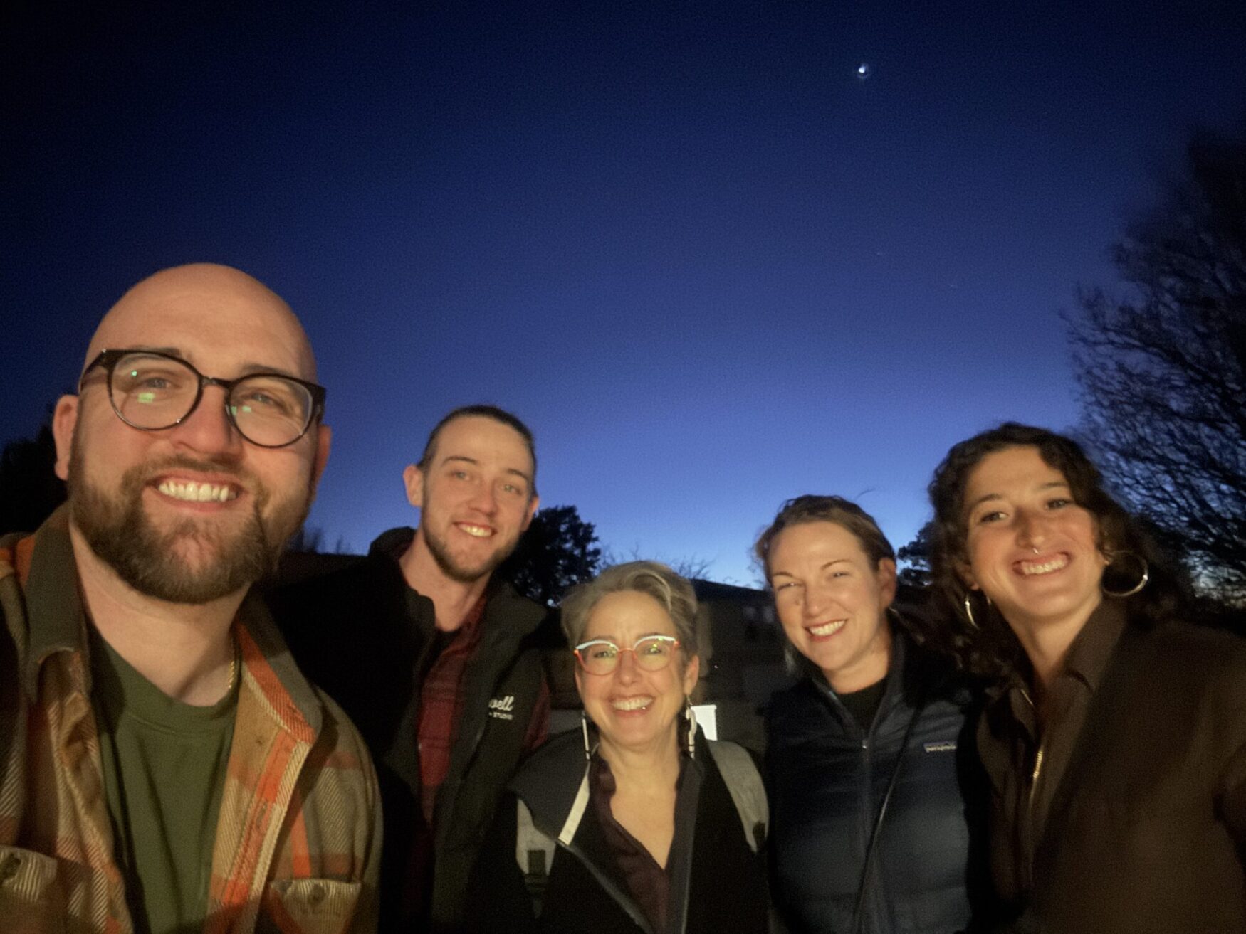 This image shows a group of five people smiling and posing for a selfie outdoors during twilight. The sky in the background is a deep blue with a visible crescent moon and some stars. The group consists of individuals dressed in warm clothing, suggesting a cool evening. They appear happy and close-knit, likely capturing a memorable moment together.