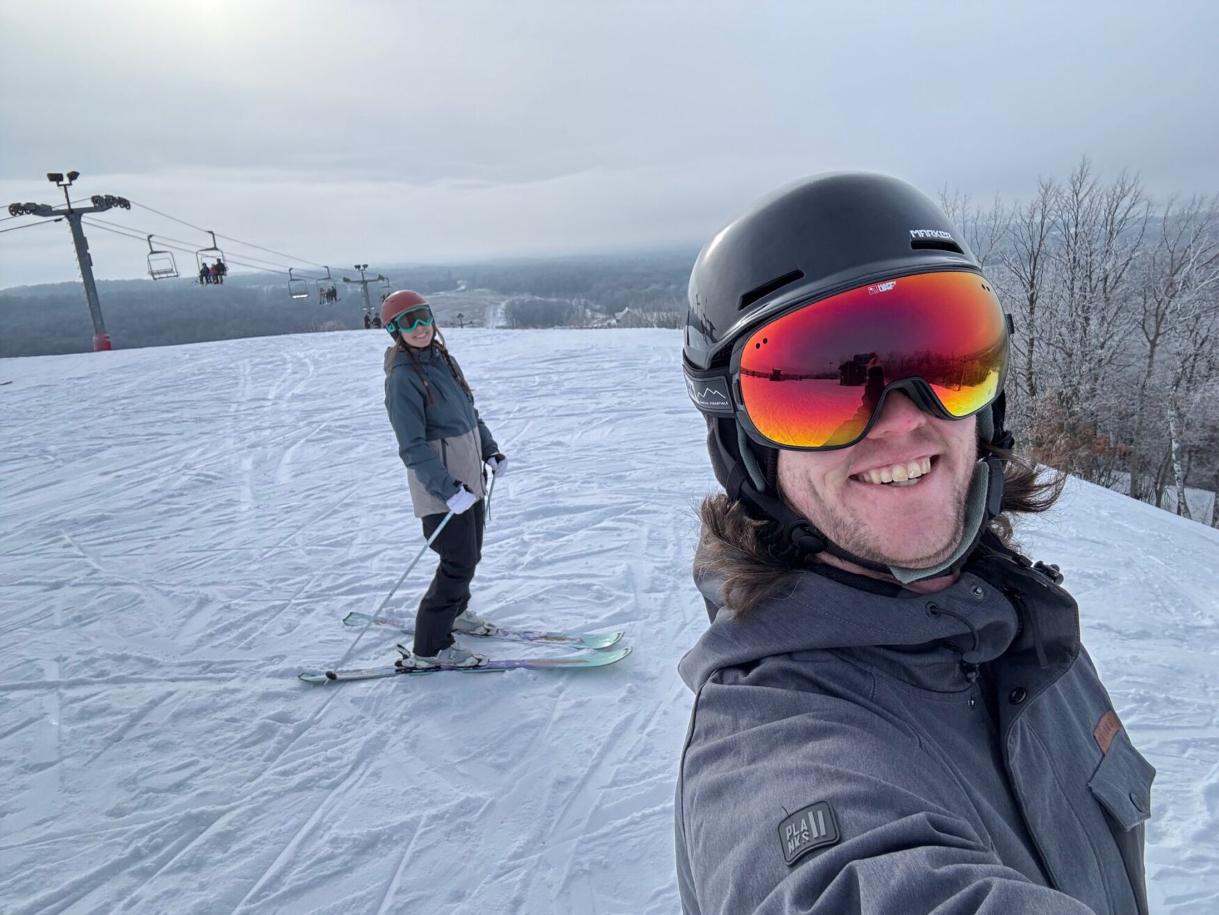 This is a fun photo of two people enjoying a day on the slopes, skiing or snowboarding in a snowy setting. It captures a cheerful moment with one person smiling at the camera in the foreground while the other is further back, standing with ski poles. The snowy landscape and ski lift in the background add to the wintery vibe.