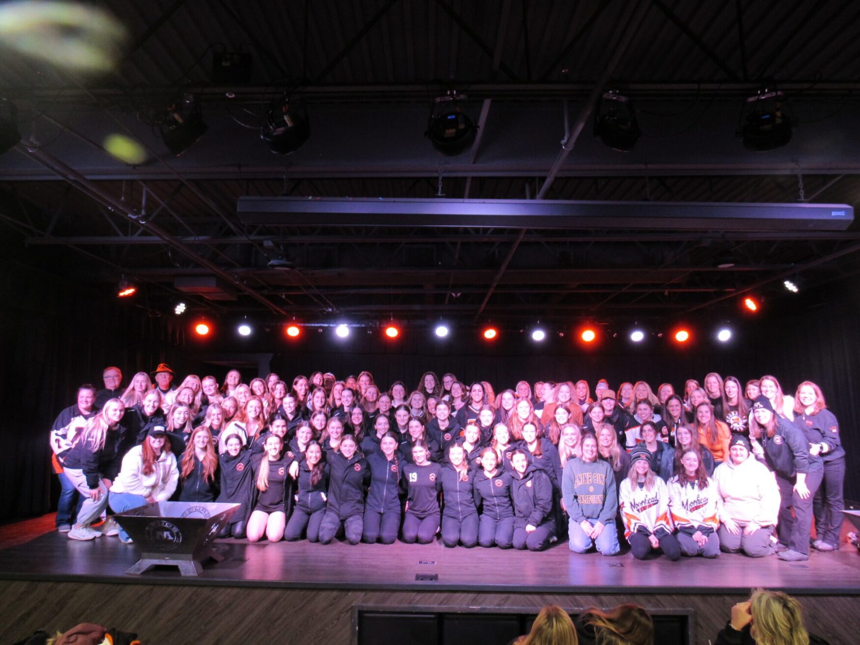 A large group photo of a sports team or organization gathered on a stage under a ceiling of industrial lighting. The group consists of over 80 individuals, predominantly women, wearing matching black athletic attire, jerseys, or team apparel. The stage is illuminated with red and white spotlights, creating a vibrant and celebratory atmosphere. Some team members kneel or sit in the front row, while others stand behind them in rows. The setting appears to be a theater or event space.