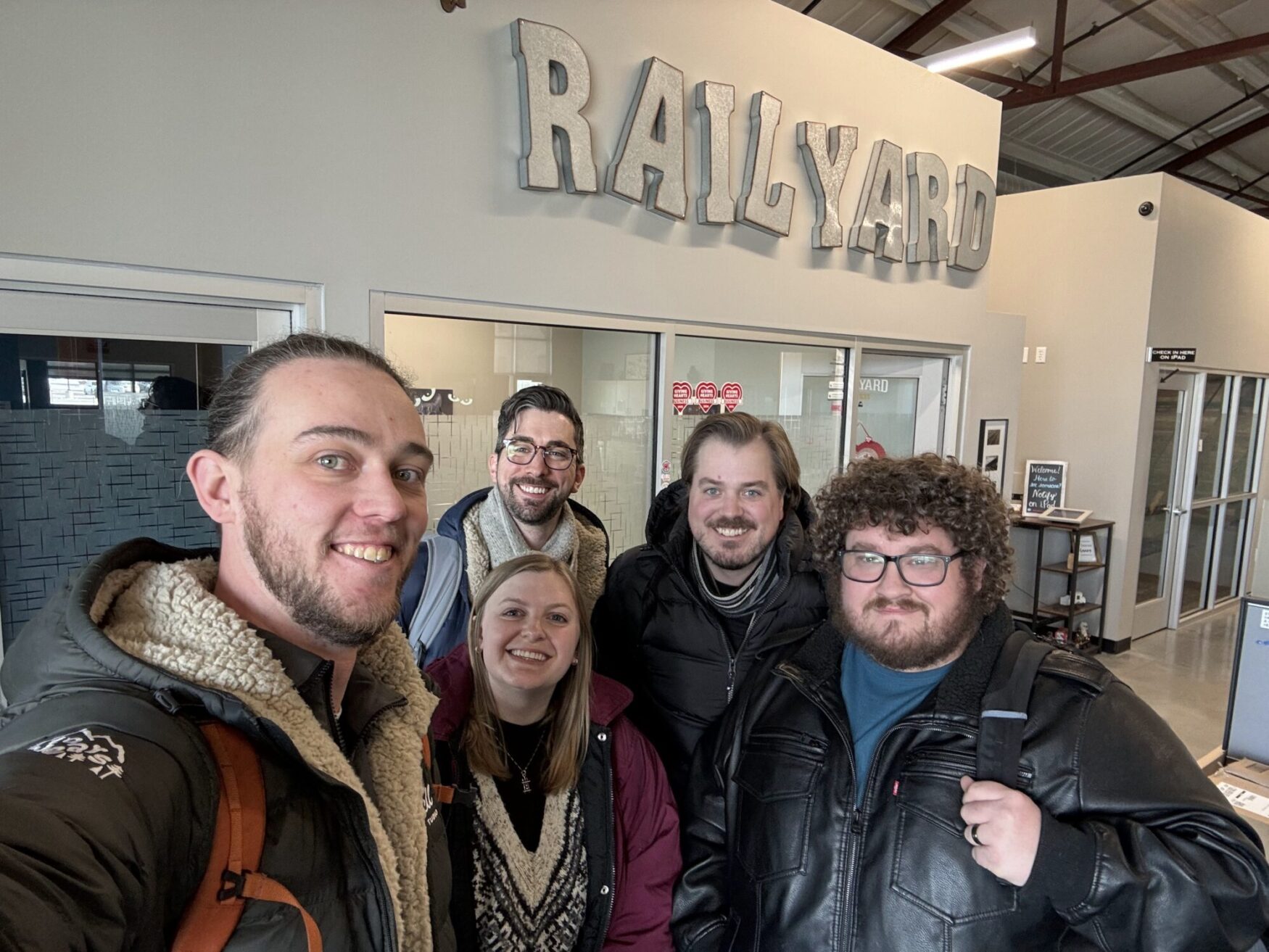 A group of five people is smiling for a selfie in front of a sign that says "RAILYARD" in large, metallic letters. They are dressed warmly in winter jackets, with a modern industrial-style space in the background featuring glass doors, exposed beams, and neutral tones. The atmosphere suggests a friendly and casual moment, possibly at a workspace or community hub.