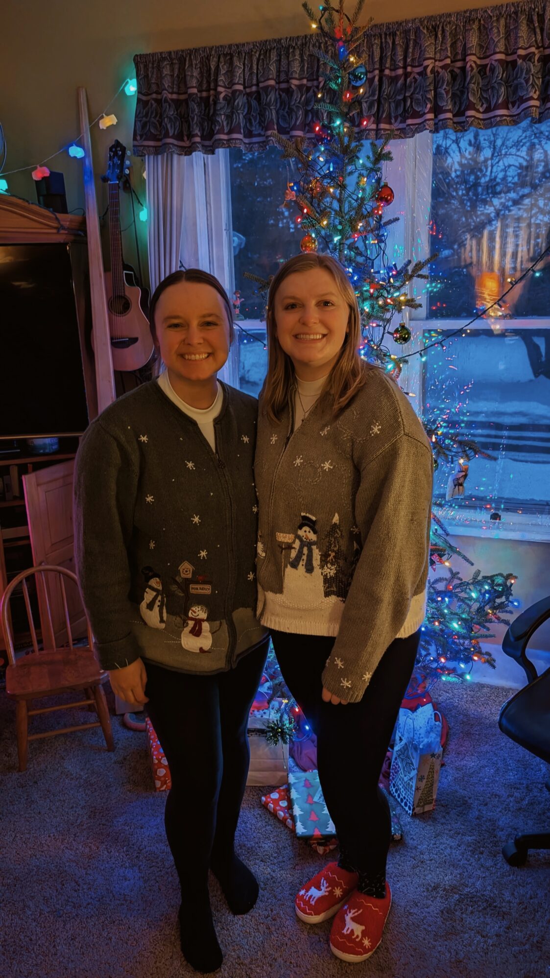 Two women pose for a photo in front of a Christmas tree. They are both wearing green sweaters with snowflakes and snowmen on them. The atmosphere is warm and festive.