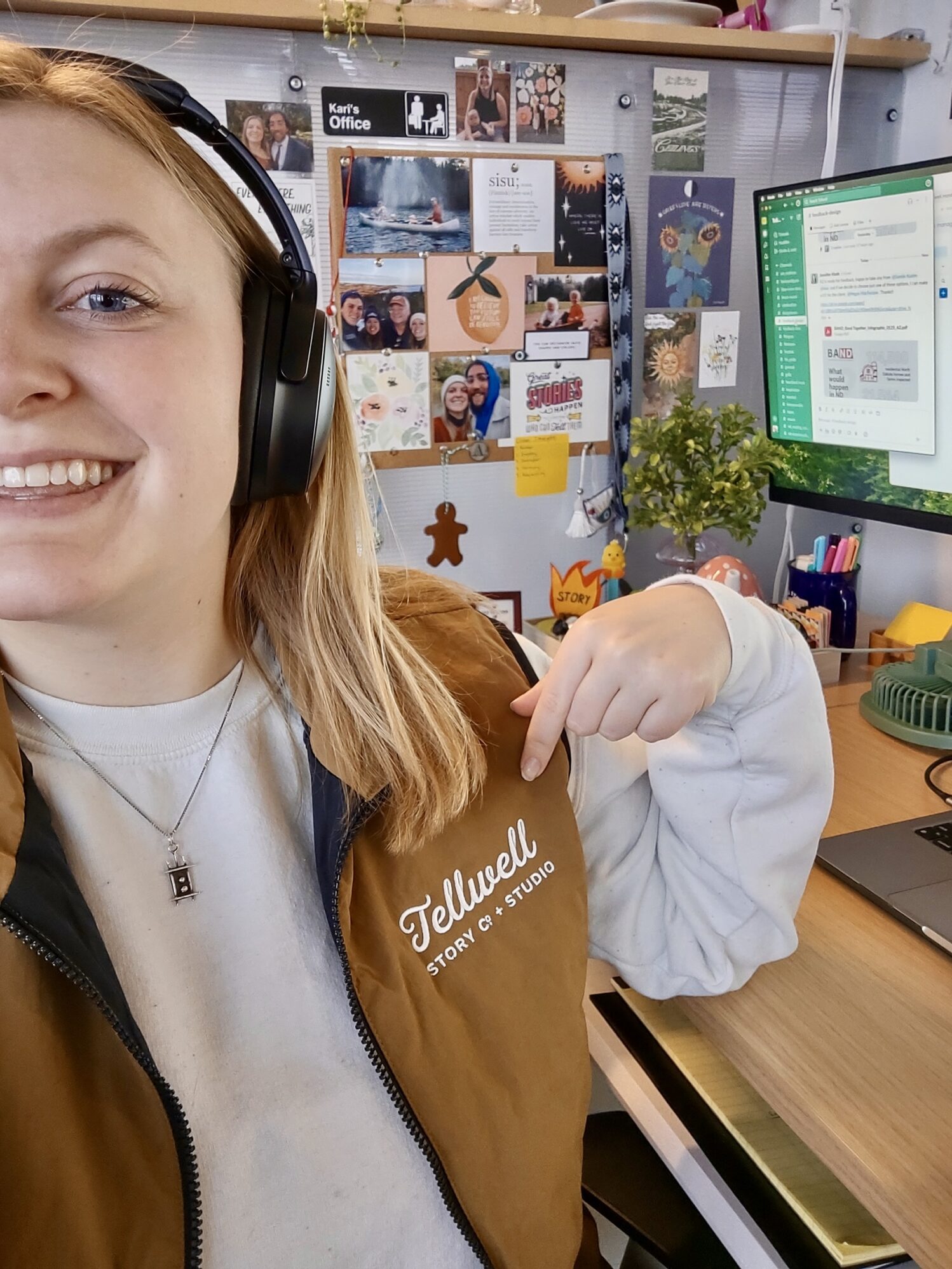 A smiling person wearing headphones points to their brown vest embroidered with the text "Tellwell Story Co. + Studio." They are sitting at a desk with a computer screen displaying Slack messages, while a pinboard full of photos, notes, and decorations is visible in the background, adding a personal touch to the workspace.
