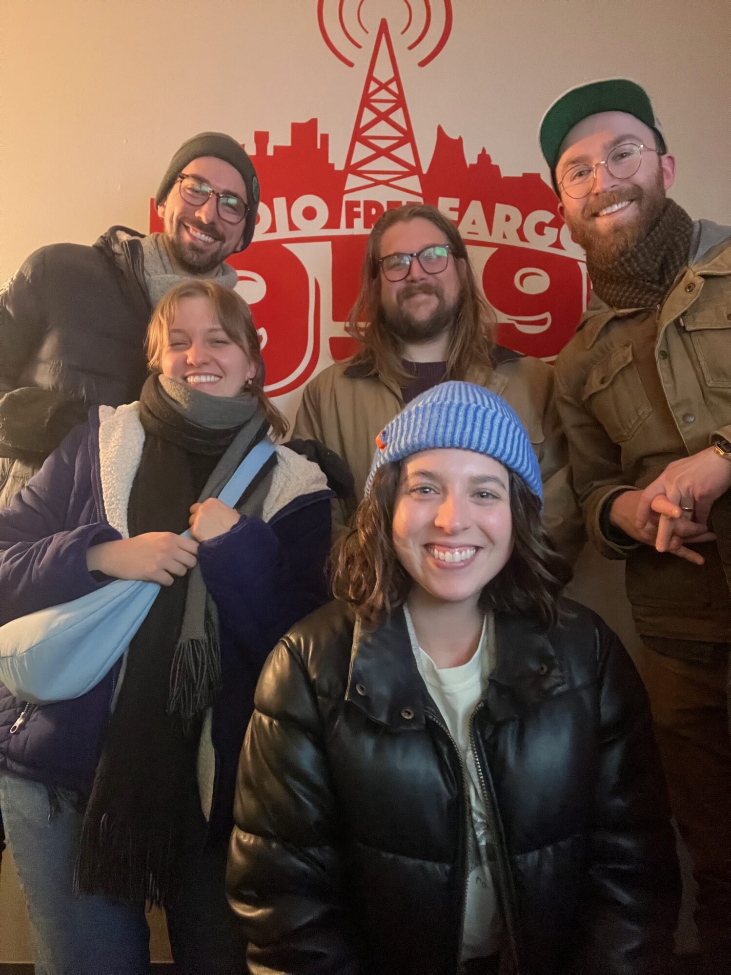 A group of five - two women and three men - pose for a photo together. The wall behind them reads “Radio Free Fargo 95.9” suggesting they are at a radio station studio. They are all dressed in winter attire, donning warm coats, winter hats, and scarfs.