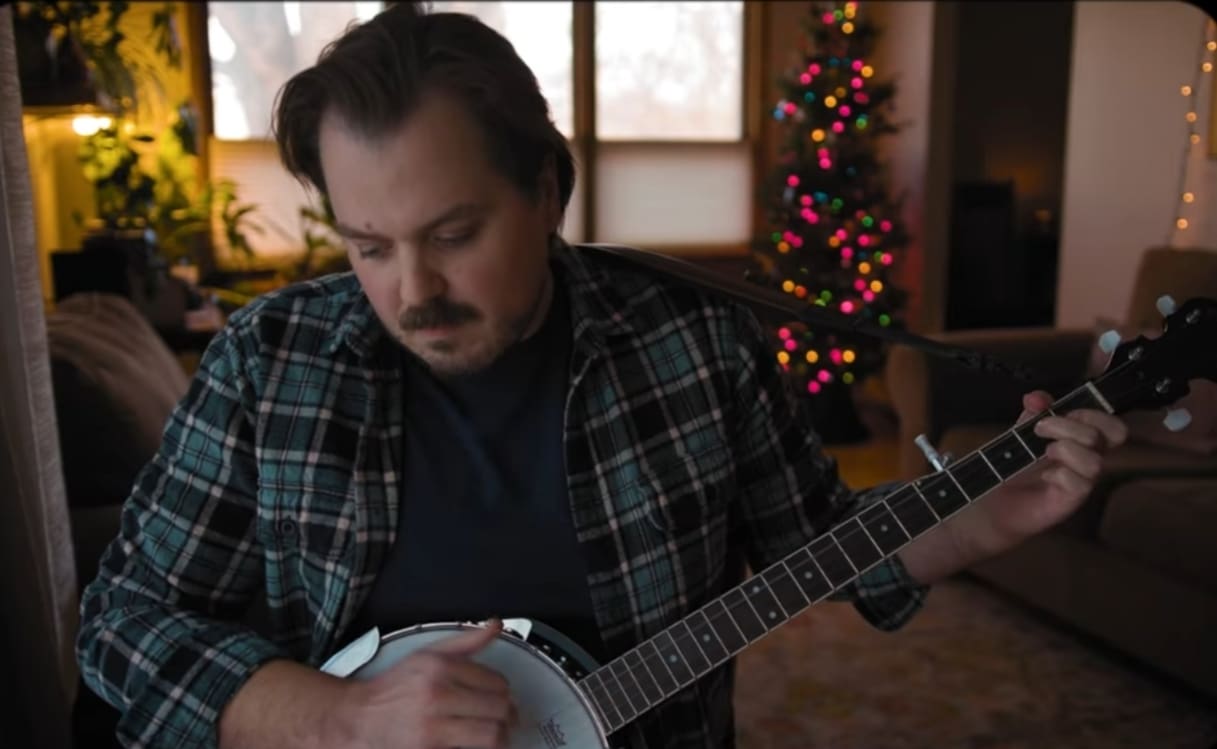 A man wearing a flannel shirt playing banjo. There is a Christmas tree in the background with additional festive lights throughout the room.