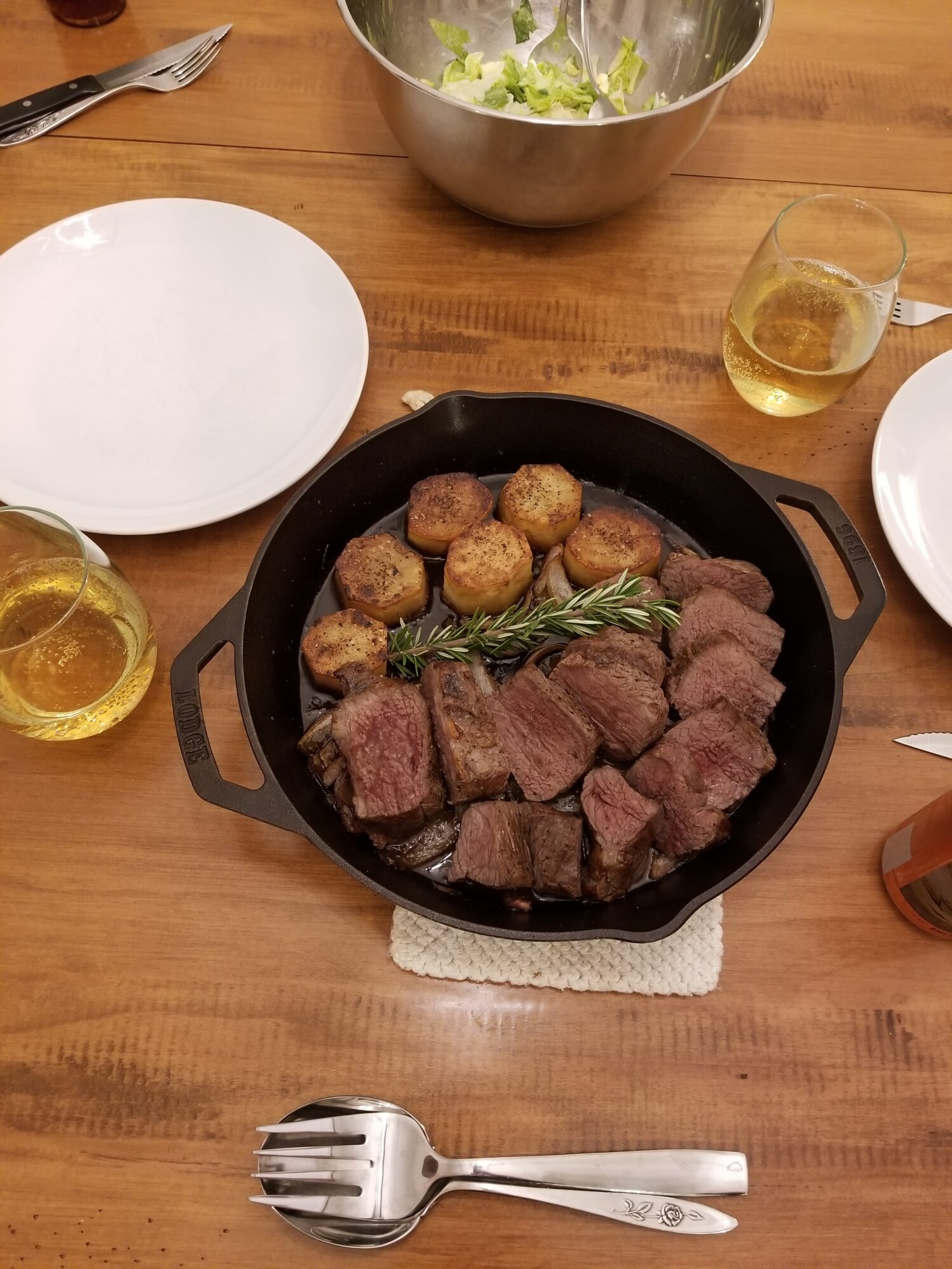 A close-up photo of a spread of food on a wooden table. There is a cast iron pan that has steak slices, potatoes, and a sprig of rosemary in it. There are two glasses of white wine and a bowl of salad along with place settings.