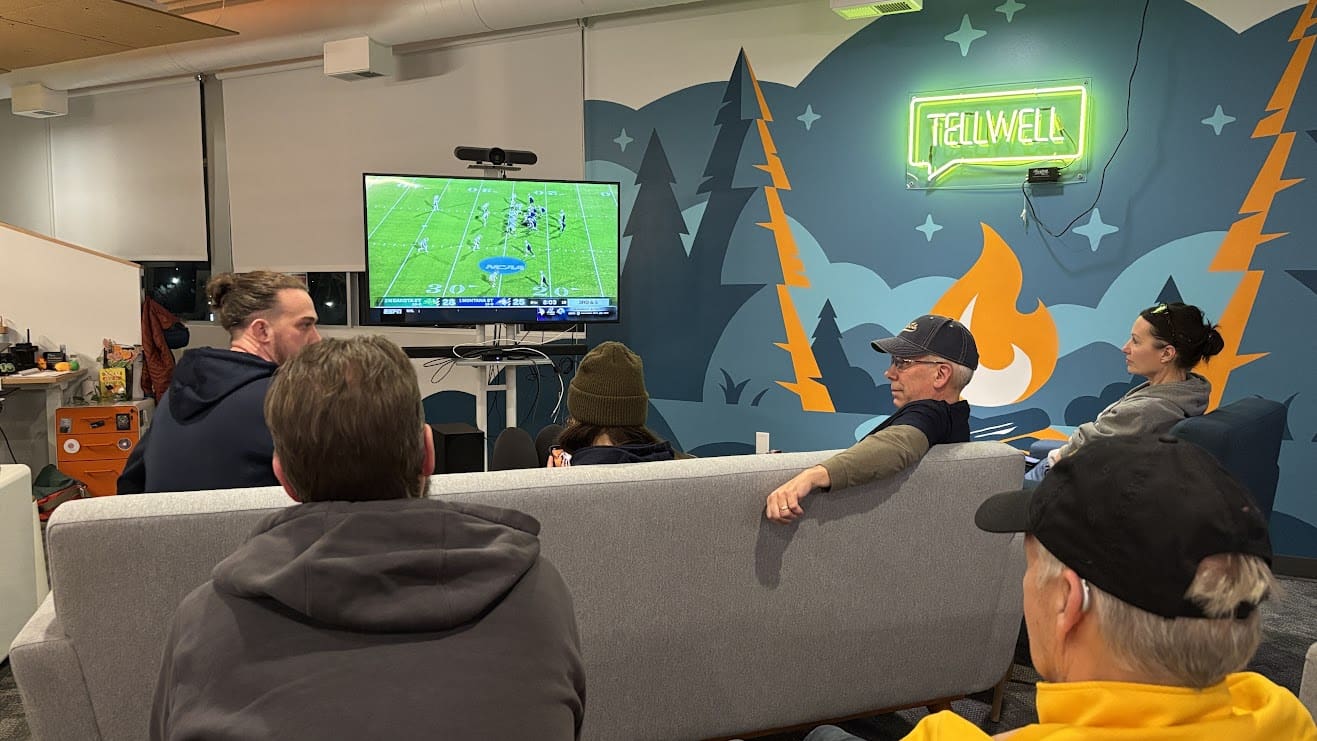A group of people gathers in a cozy, modern space, watching a football game on a large TV. They sit on a gray couch with a mural of a campfire and trees behind them, illuminated by a neon green "Tellwell" sign. The atmosphere is casual and relaxed, with warm tones and a touch of outdoor-inspired decor.