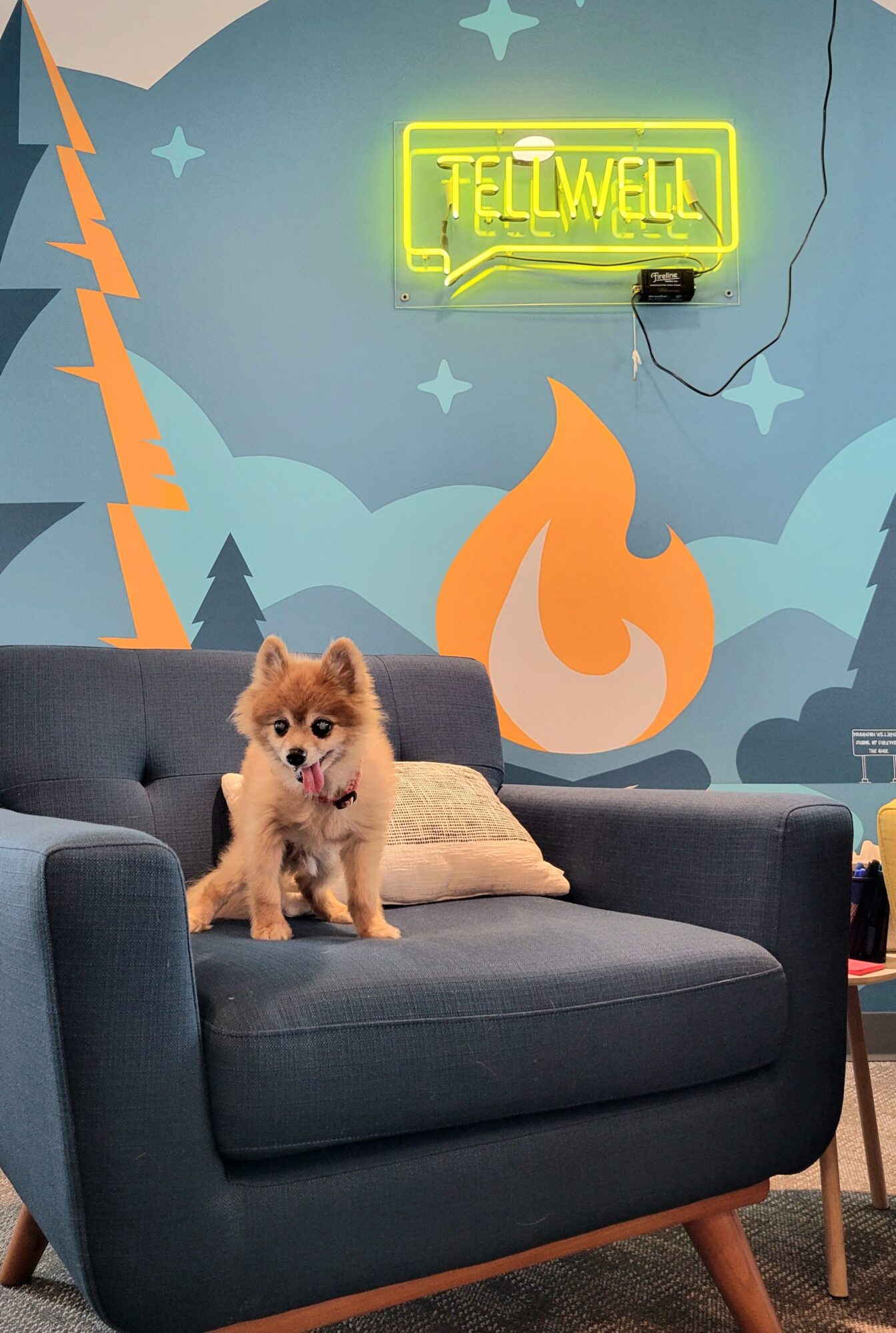 A small, fluffy Pomeranian with a reddish-brown coat and a slightly protruding tongue sits on a modern blue armchair. Behind the dog, a colorful mural with a camping theme features stylized mountains, trees, stars, and a campfire. A neon yellow "TELLWELL" sign glows on the wall, with its power cord visible. The cozy scene is set in a creative and inviting workspace.