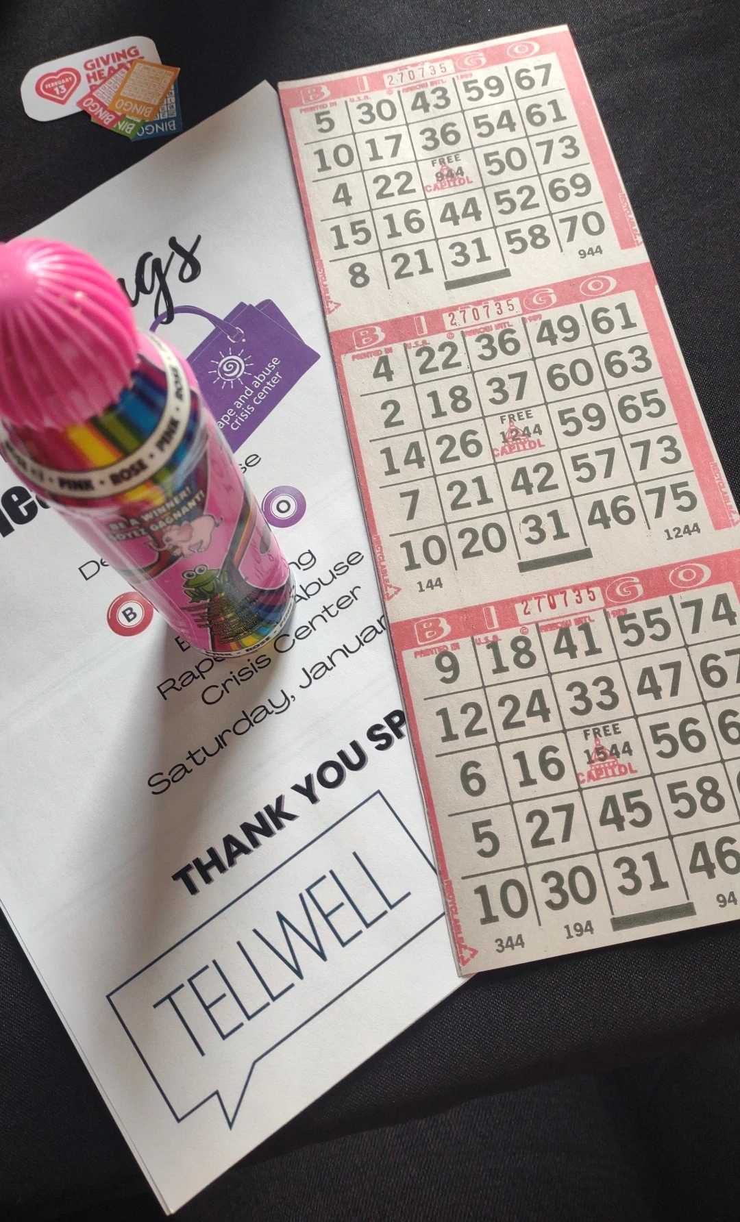 A close-up of bingo cards, a pink bingo dauber featuring a cartoon frog, and an event program on a black surface. The program mentions a fundraiser for the Rape and Abuse Crisis Center and includes a "THANK YOU SPONSOR" section with the Tellwell logo. A small "Giving Hearts Day" sticker is also visible.