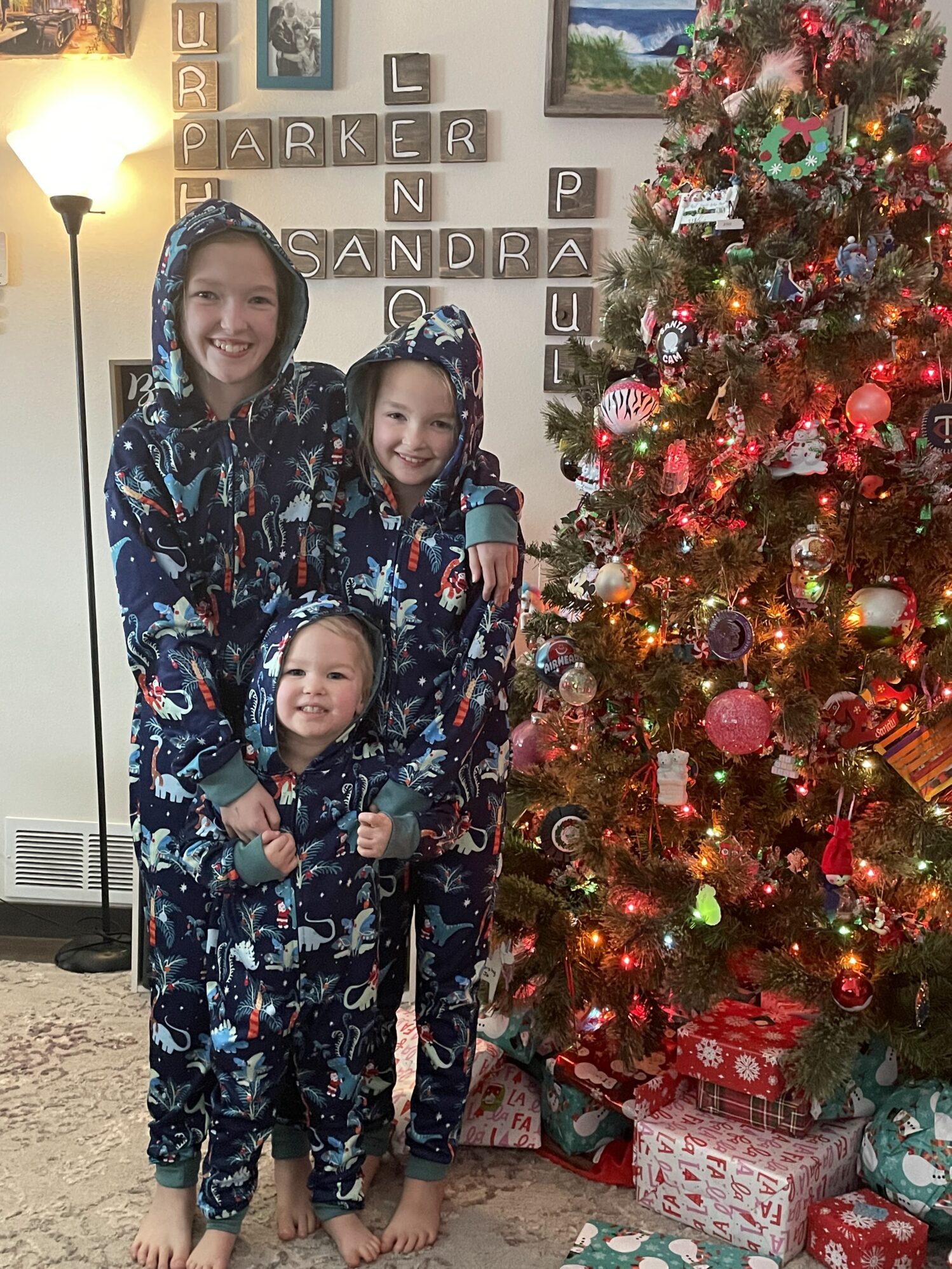 Three girls stand next to a Christmas tree wearing matching pajamas. They are smiling and the atmosphere is happy and festive. There are gifts wrapped under the tree.