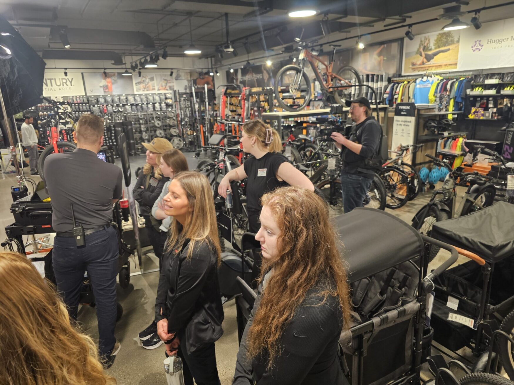 This image shows a group of people inside a well-stocked outdoor or sporting goods store. The store contains various bicycles, bike accessories, and other fitness-related gear. Some individuals are engaged in conversation, while others appear to be involved in a filming or production process, as evidenced by professional camera equipment and lighting. The atmosphere suggests a business or promotional event, possibly related to cycling or outdoor activities.