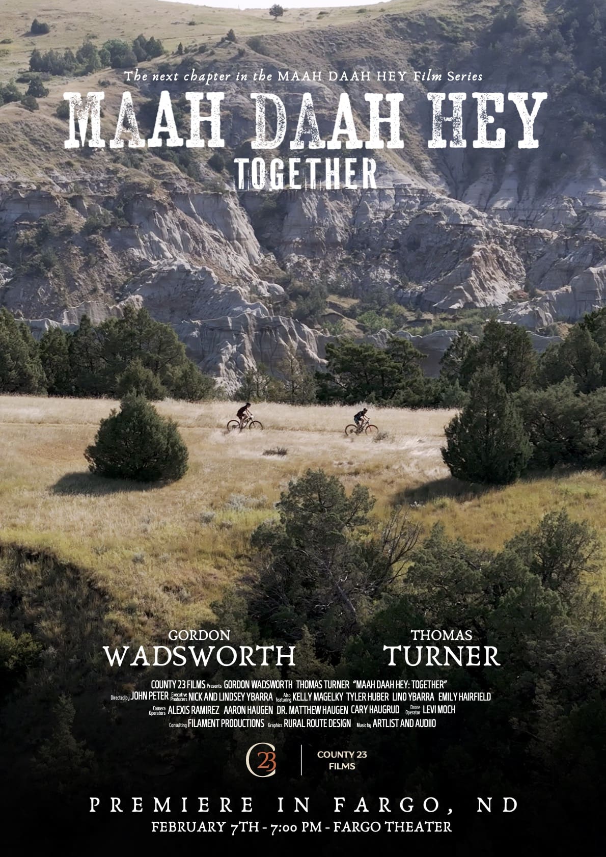 A promotional poster for the film Maah Daah Hey: Together, part of the Maah Daah Hey film series. The background features two cyclists riding through the rugged Badlands landscape with rolling grasslands, trees, and dramatic rock formations. The title is displayed in bold, distressed lettering at the top, with the names of featured riders Gordon Wadsworth and Thomas Turner below. Additional credits list filmmakers, sponsors, and production teams. The bottom announces the film’s premiere in Fargo, ND, on February 7th at 7:00 PM at the Fargo Theatre.